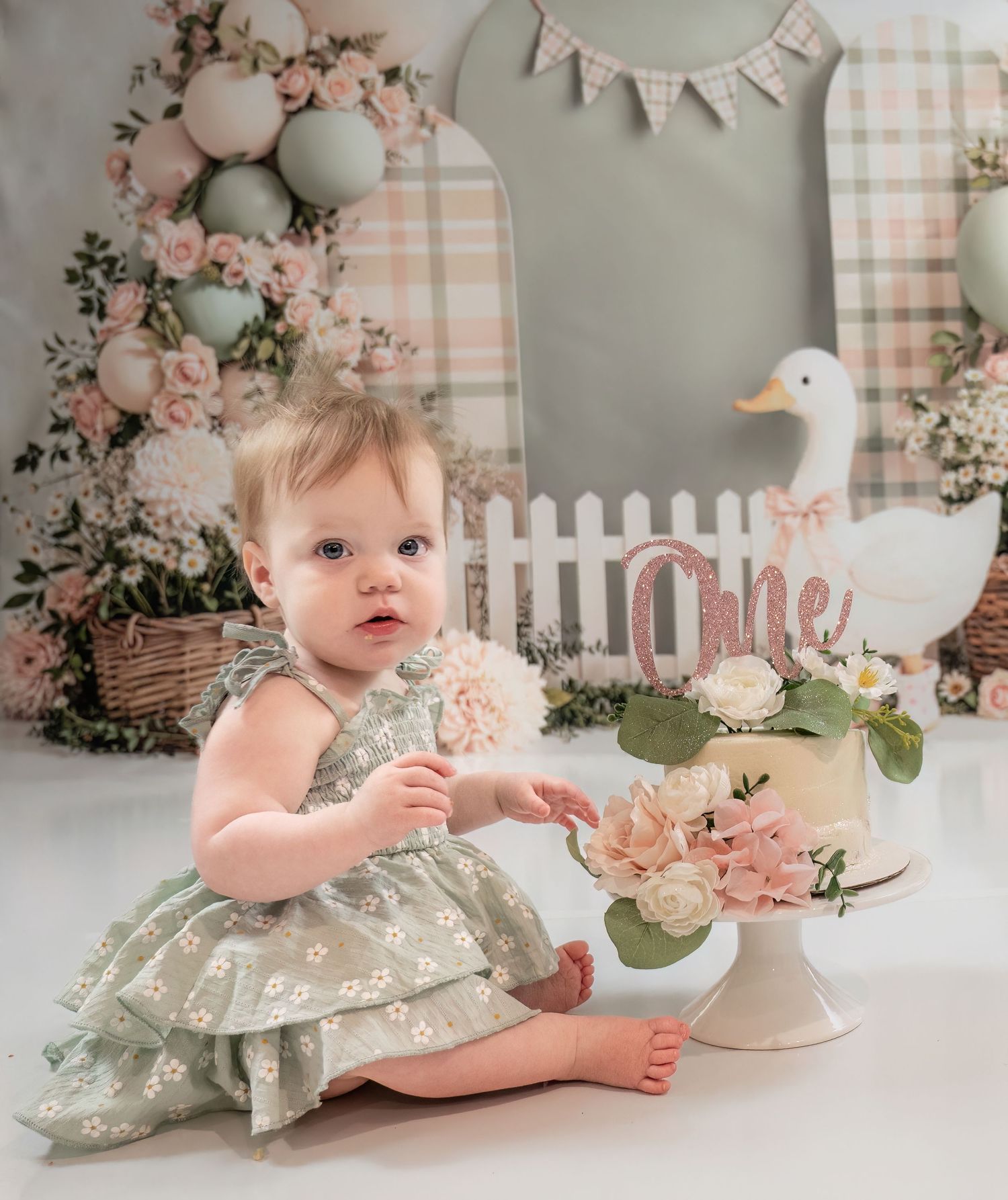 Baby girl in floral sage dress sits beside rose-decorated first birthday cake at duck-themed cake smash session.
