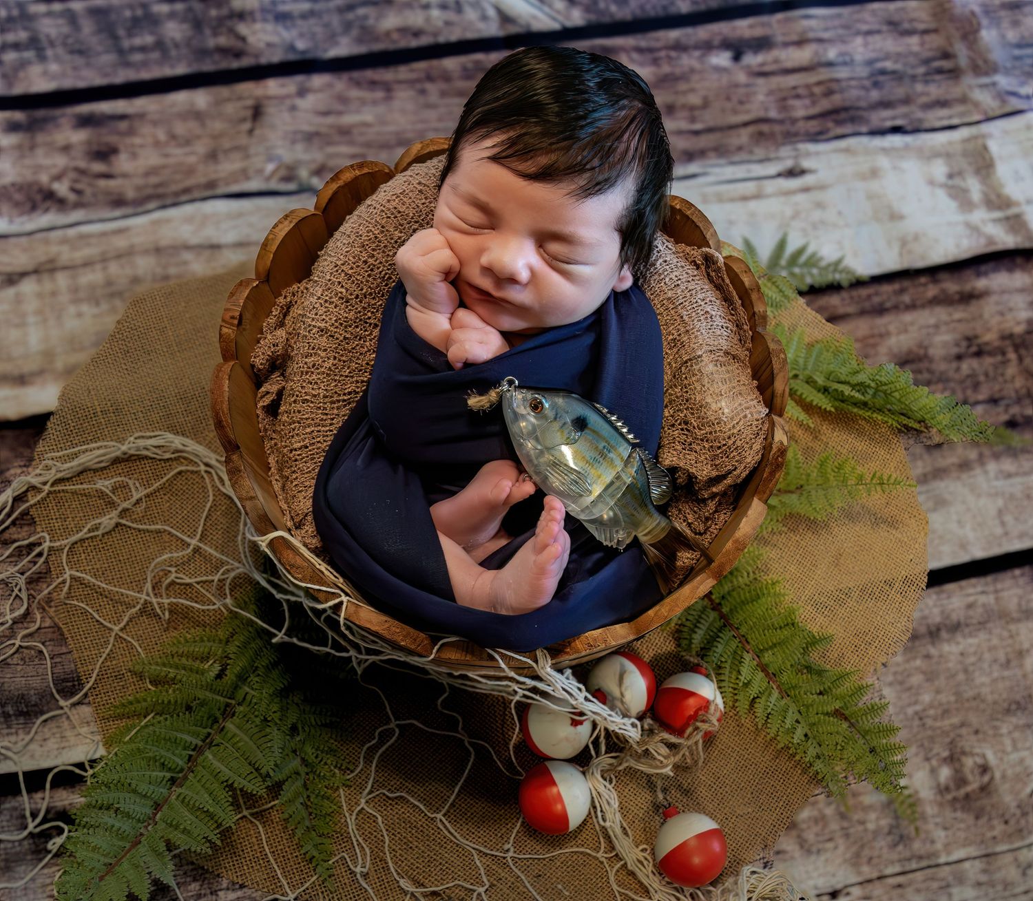 Smiling newborn baby posed in rustic basket with fishing bobbers and fern leaves decor.