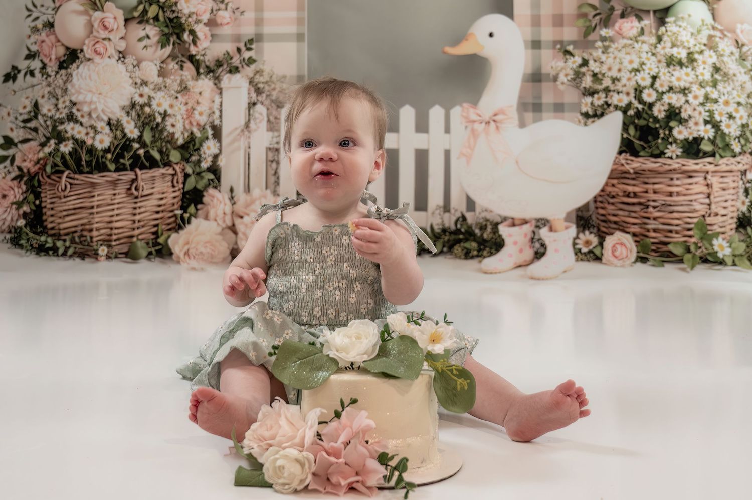 Smiling baby girl in sage romper sits before floral cake at duck-themed first birthday cake smash session.