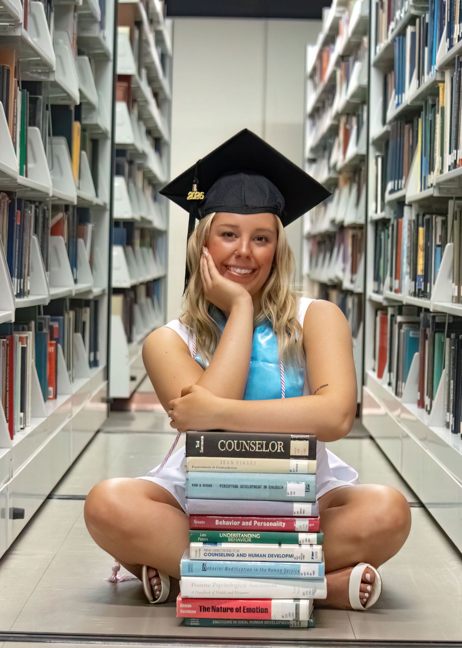 A smiling blonde graduate in a black cap poses with a stack of counseling textbooks in a library aisle.