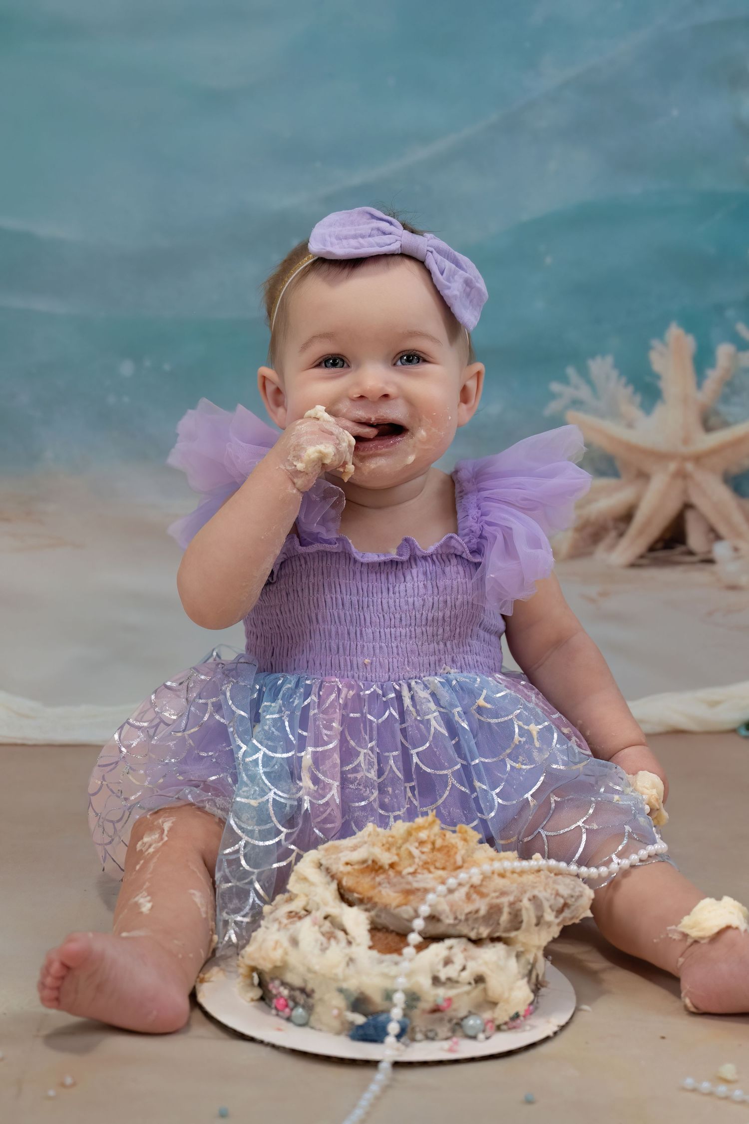 Baby girl in purple mermaid dress enjoying her first birthday cake smash.