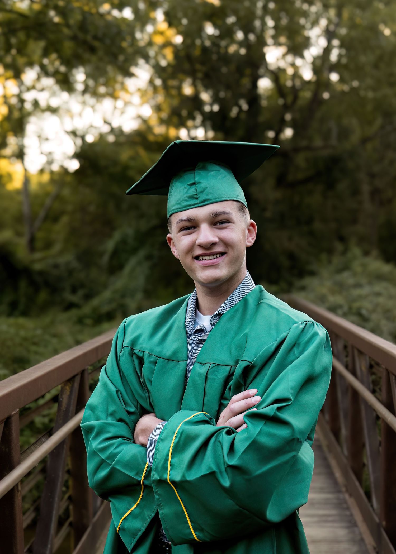 A smiling young male graduate in a green cap and gown stands with arms crossed on a wooden bridge.