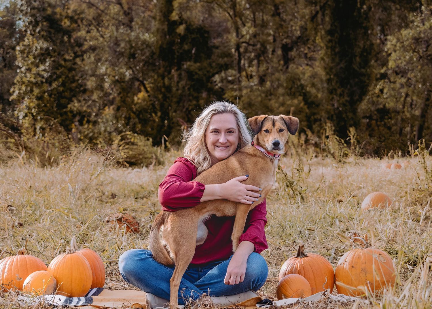 Smiling blonde woman hugging brown dog outdoors surrounded by orange pumpkins in autumn field.