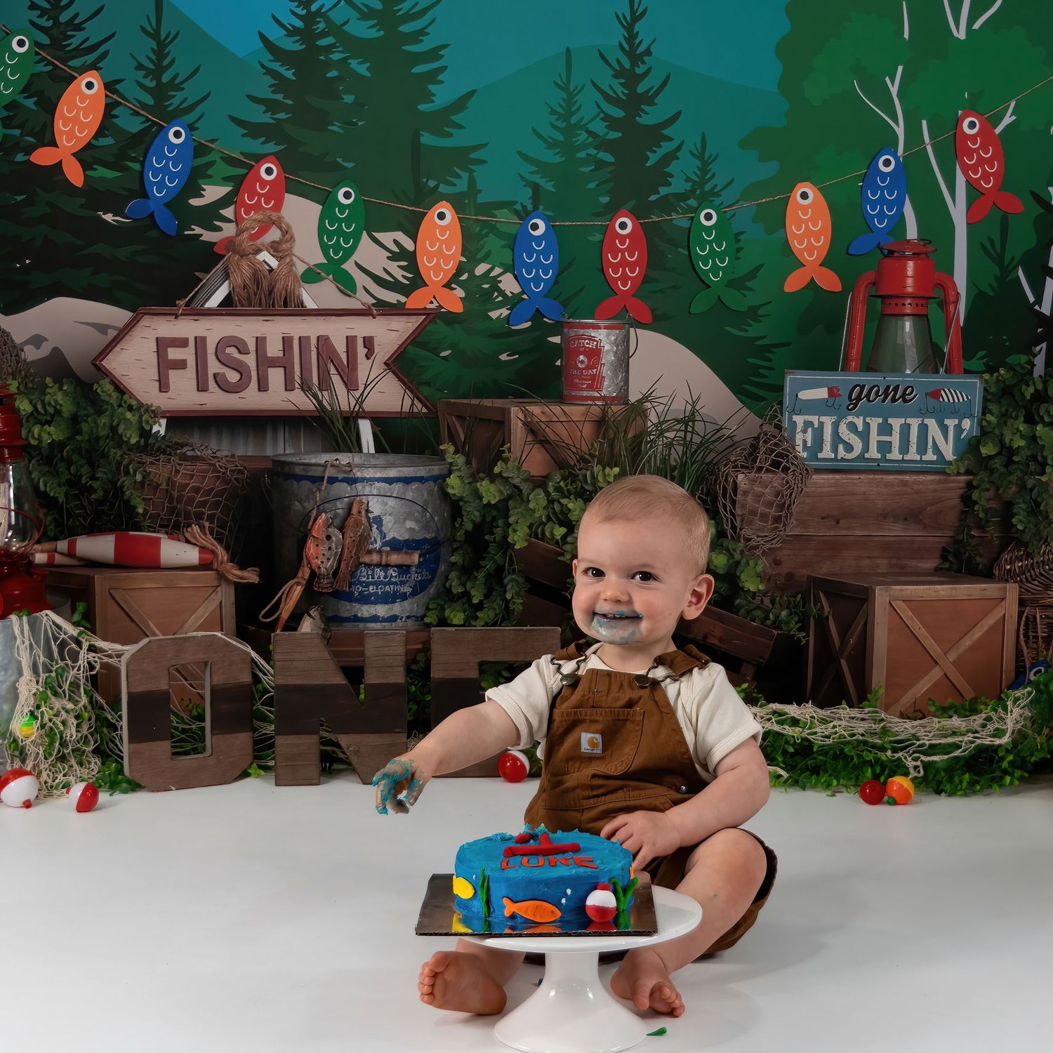 Baby boy in overalls smiles beside fishing-themed smash cake at Gone Fishin first birthday photo session.
