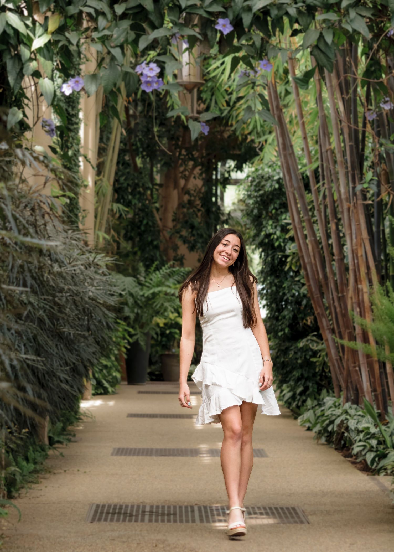 Young woman in white ruffle dress walking through lush tropical garden pathway with purple flowers.
