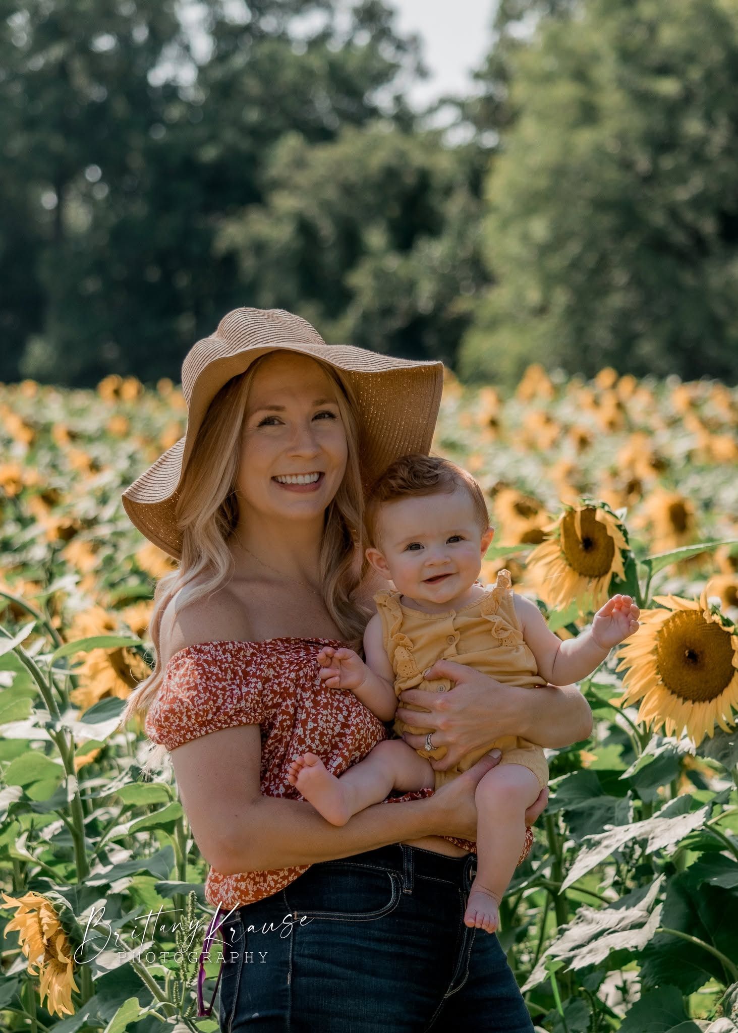 Smiling blonde woman holding a baby in a sunflower field on a sunny day.