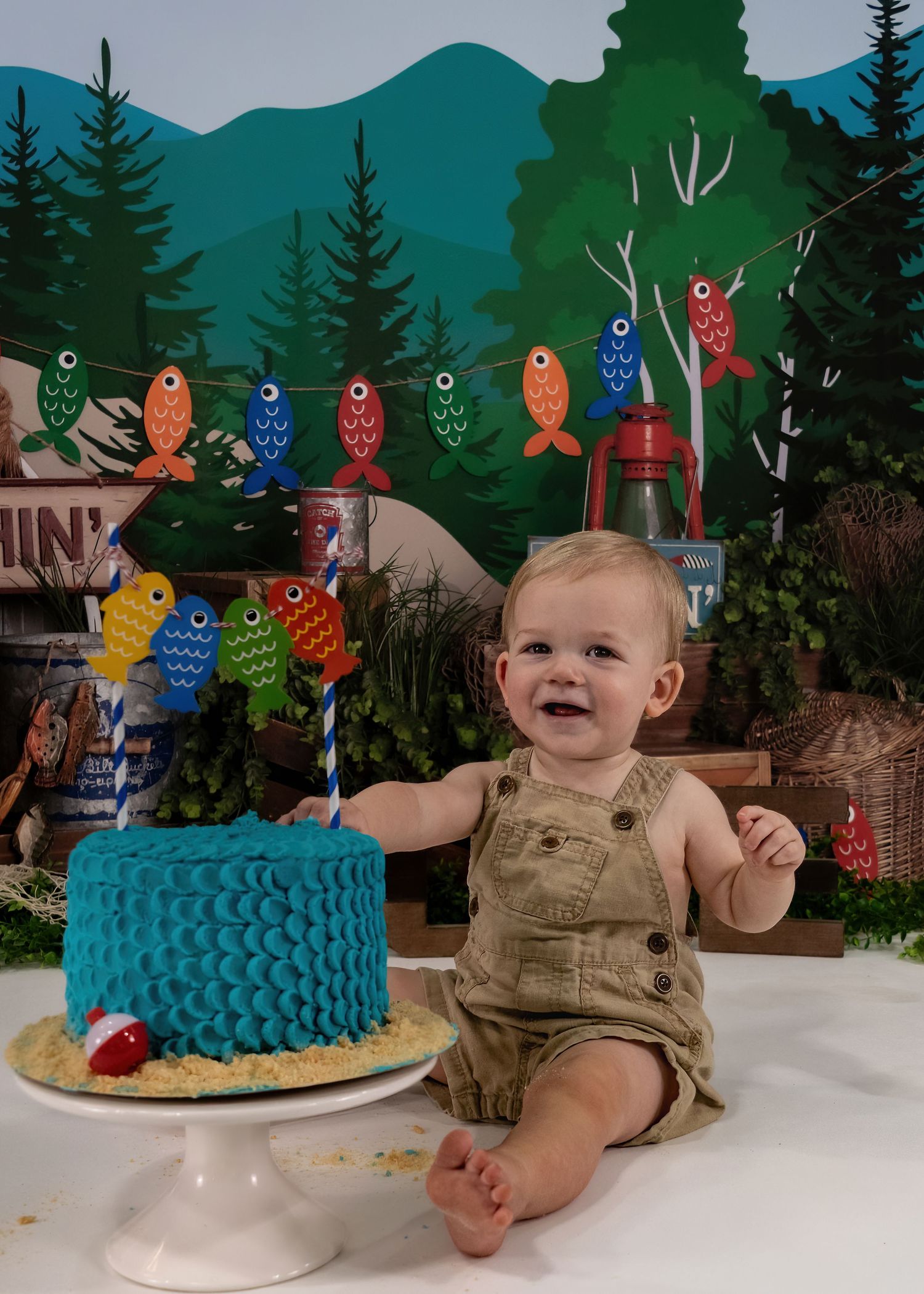 Happy baby in overalls sits beside blue fish-scale cake at colorful fishing-themed first birthday smash session.