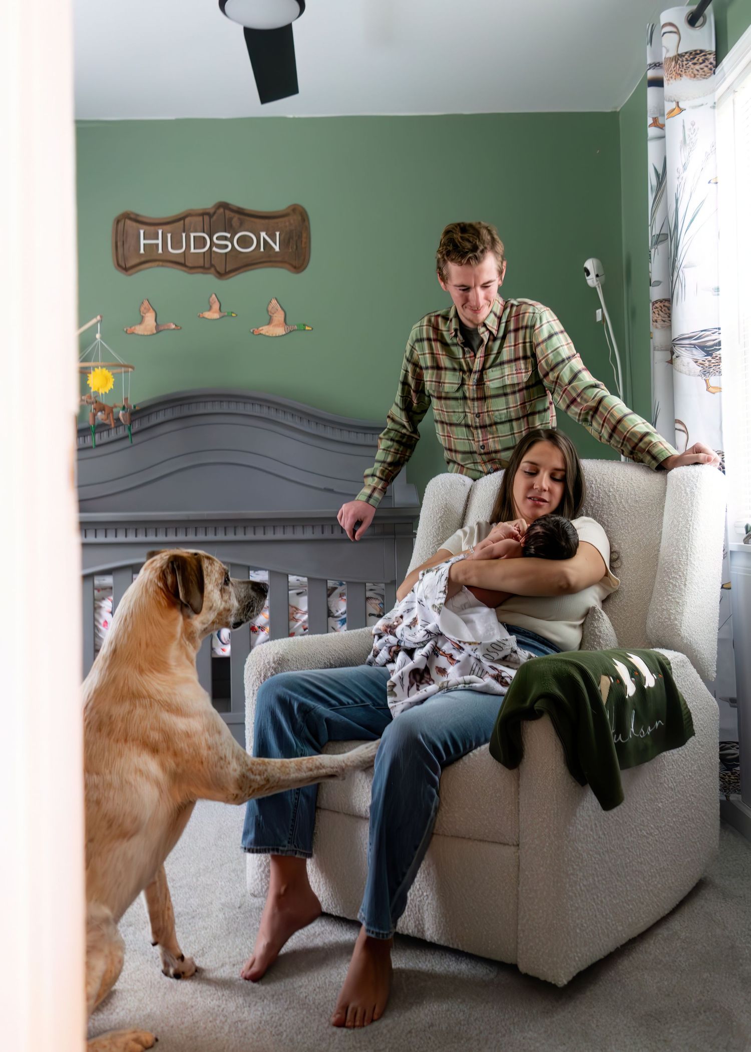 New parents with dog admiring newborn baby Hudson in green nursery with personalized name sign.