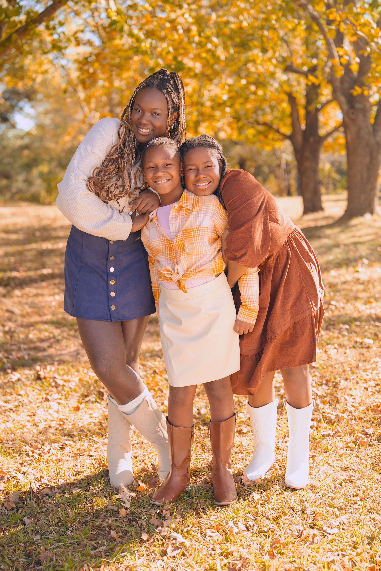Three friends embrace and laugh together on a sunny autumn day with golden fall foliage in the background.