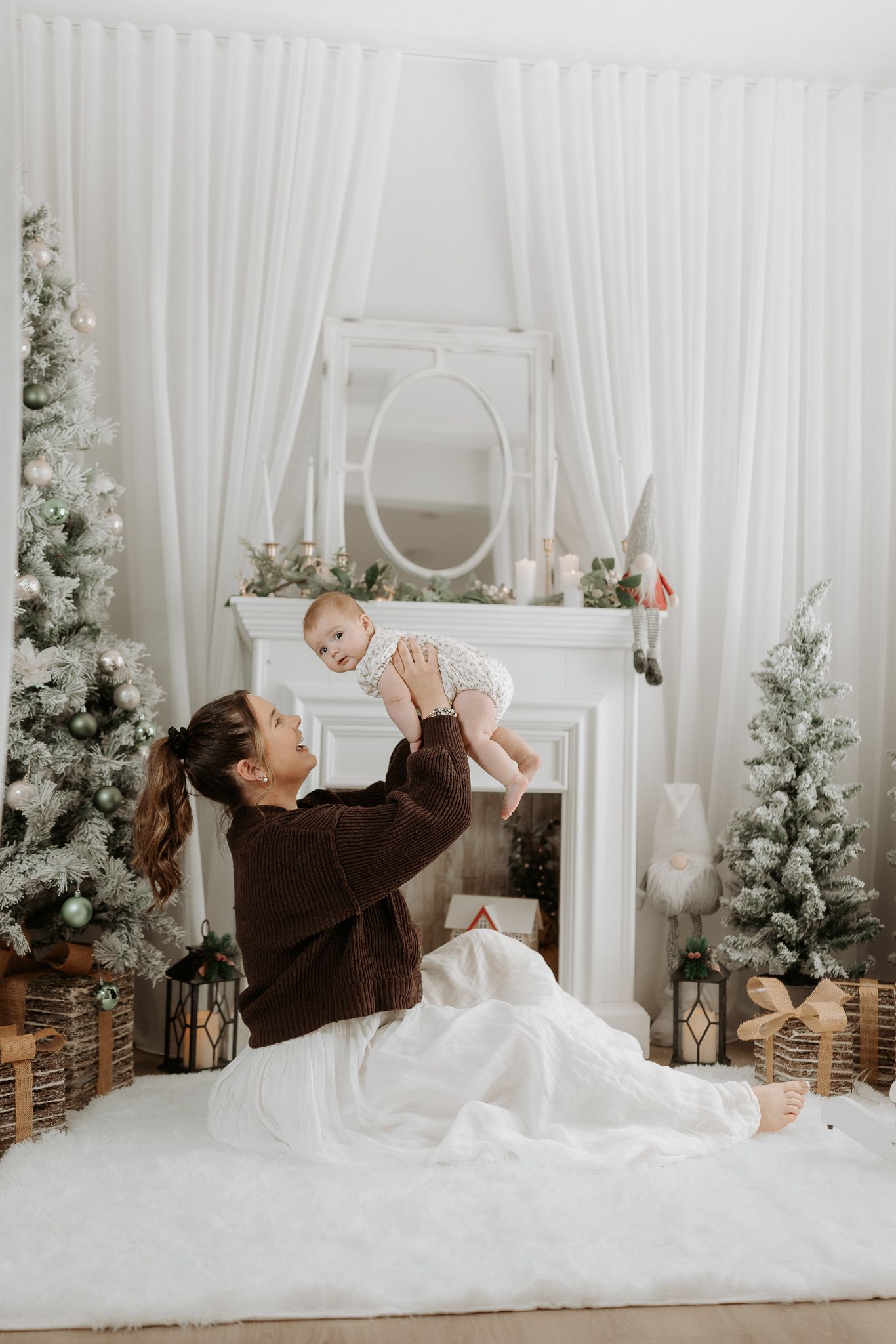 Mother lifting baby in festive white living room with Christmas trees and fireplace decor.