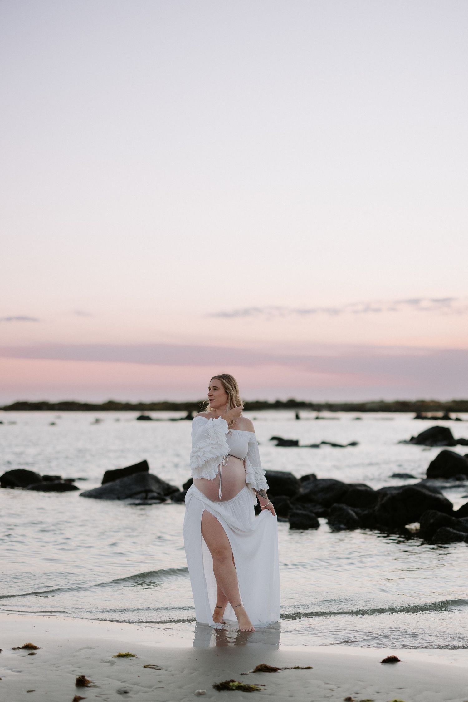 Woman in flowing white dress standing on rocky beach at sunset with pink sky.
