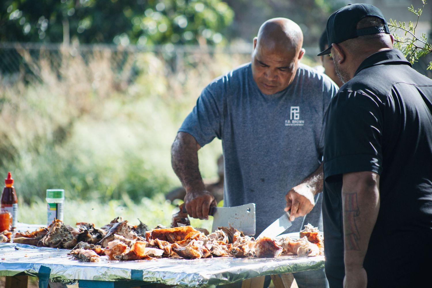 Two people preparing and carving meat at an outdoor barbecue table during a sunny day.