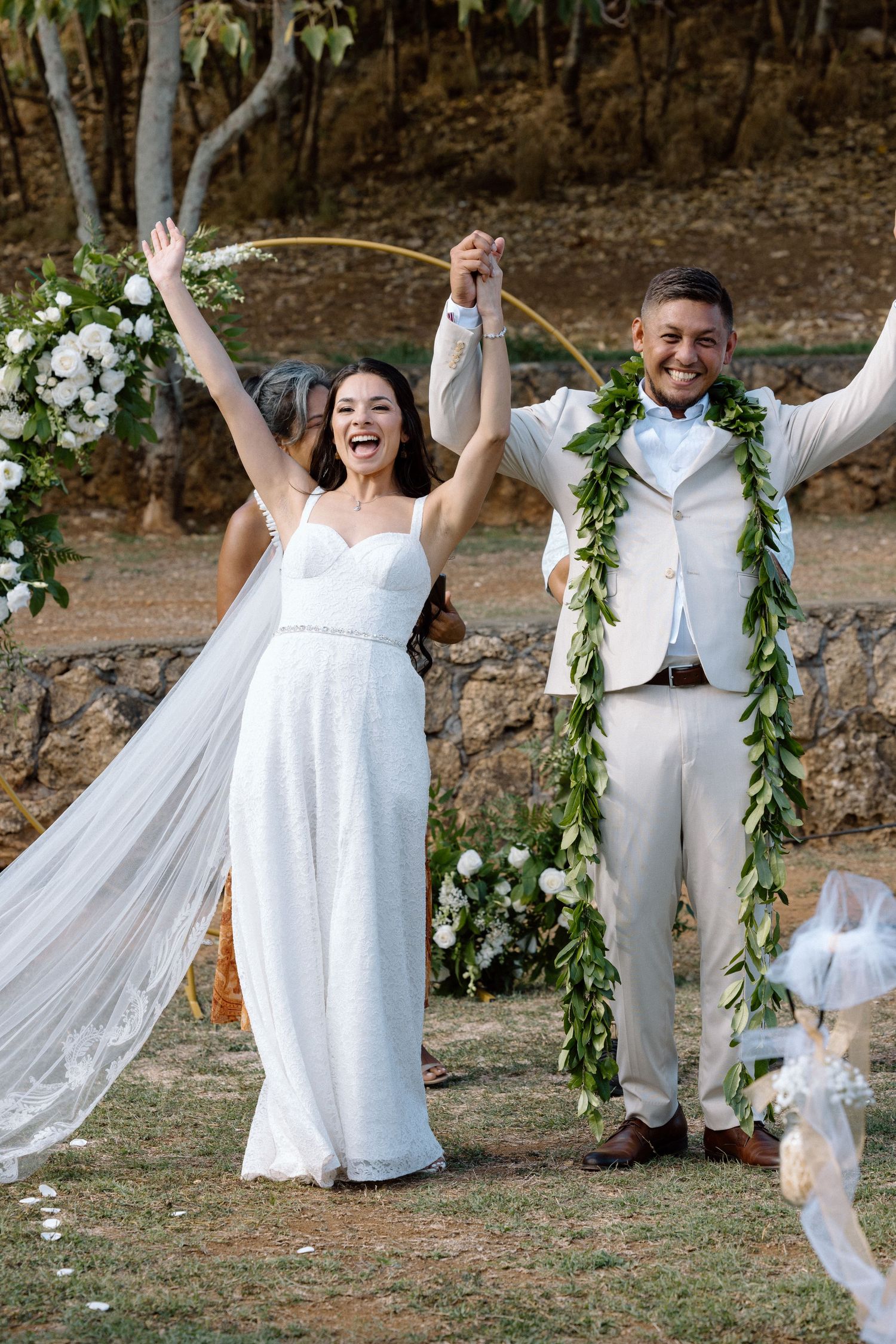 Newlyweds celebrate with raised hands under floral arch during outdoor wedding ceremony.