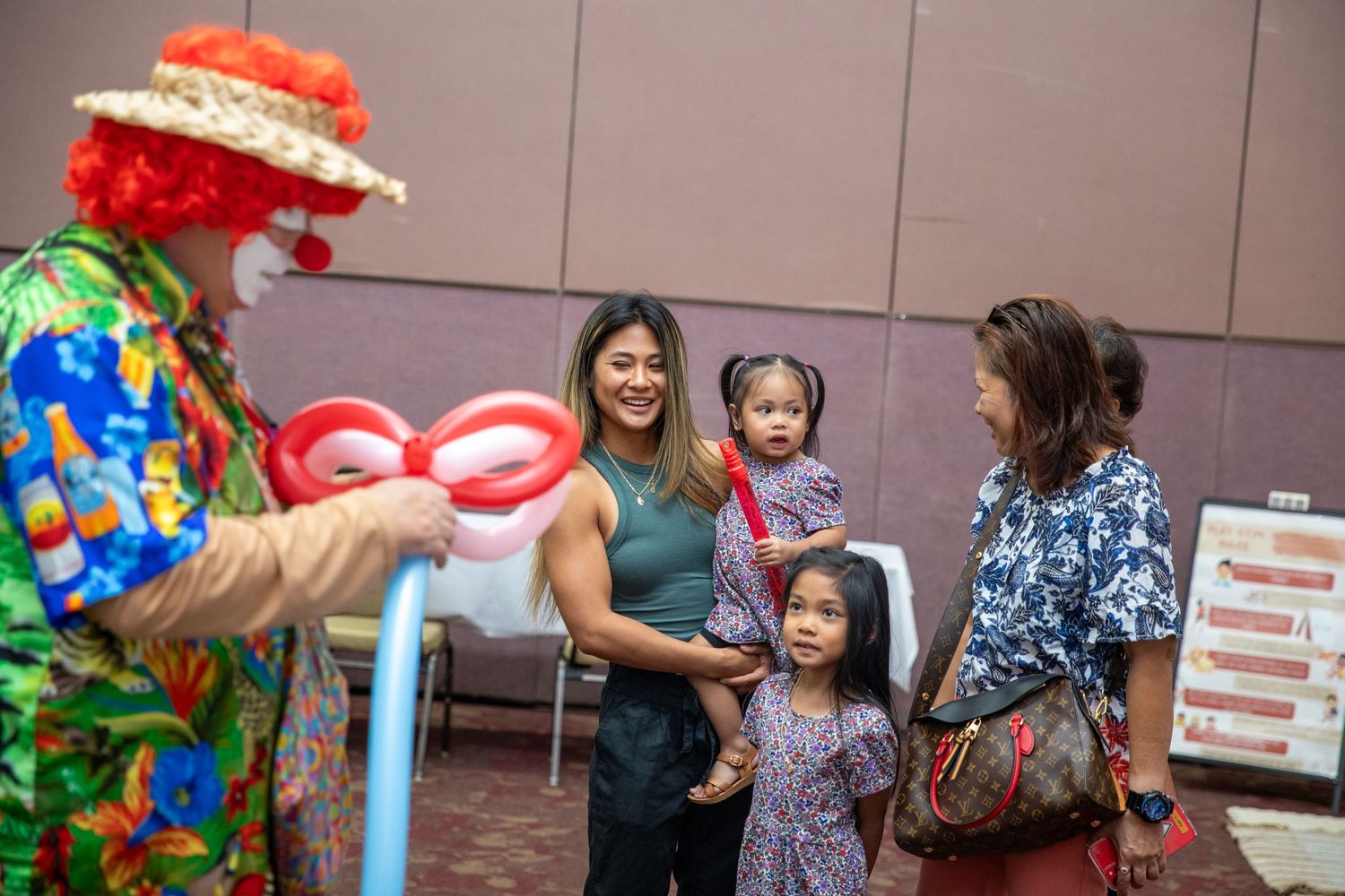 A colorfully dressed clown entertains a small group with balloon art at an indoor event.
