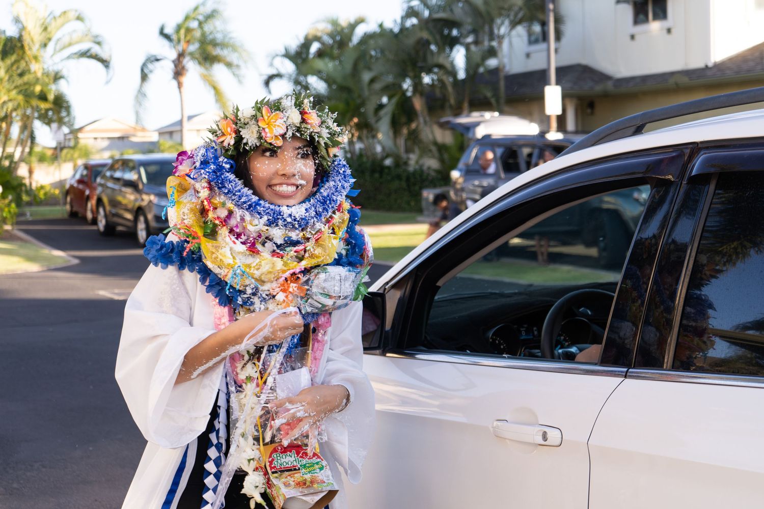 A graduate wearing colorful leis stands next to a car during a drive-by celebration.