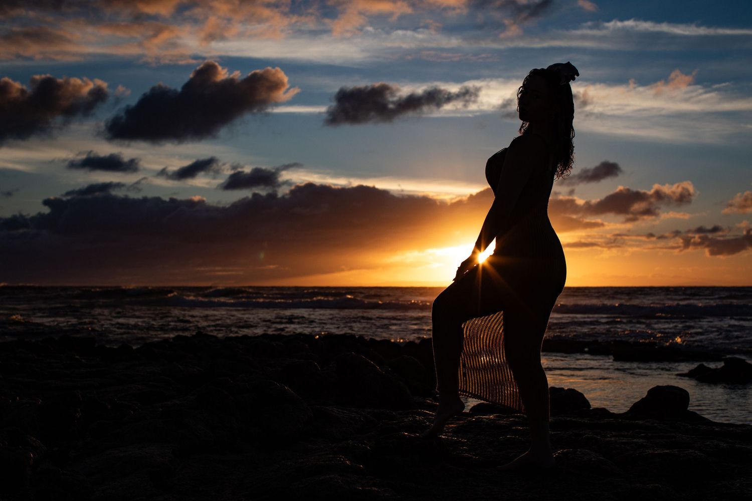 A silhouetted figure strikes a pose on rocky shore during dramatic orange sunset over the ocean.
