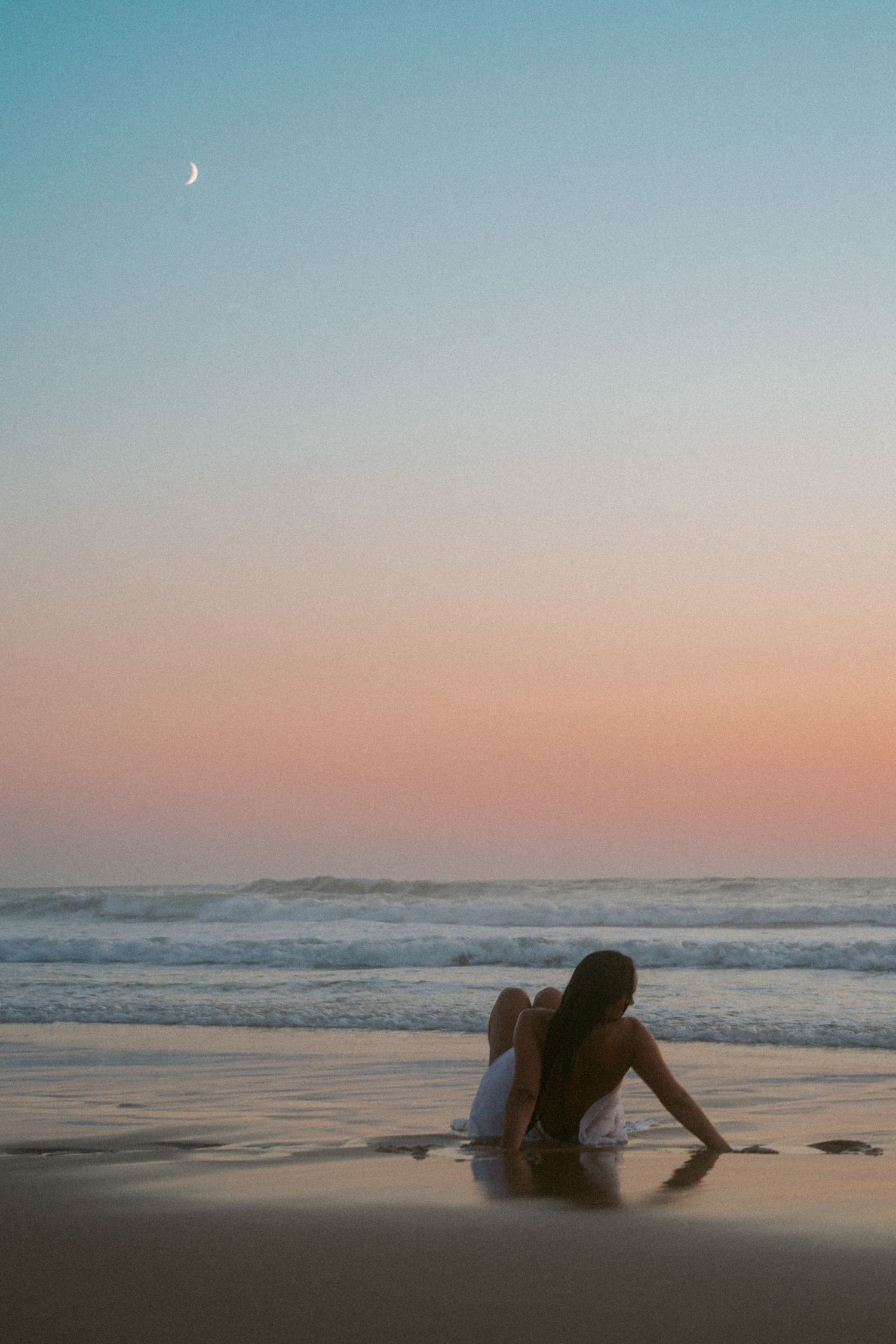 Silhouette sitting on beach at sunset with crescent moon in sky.