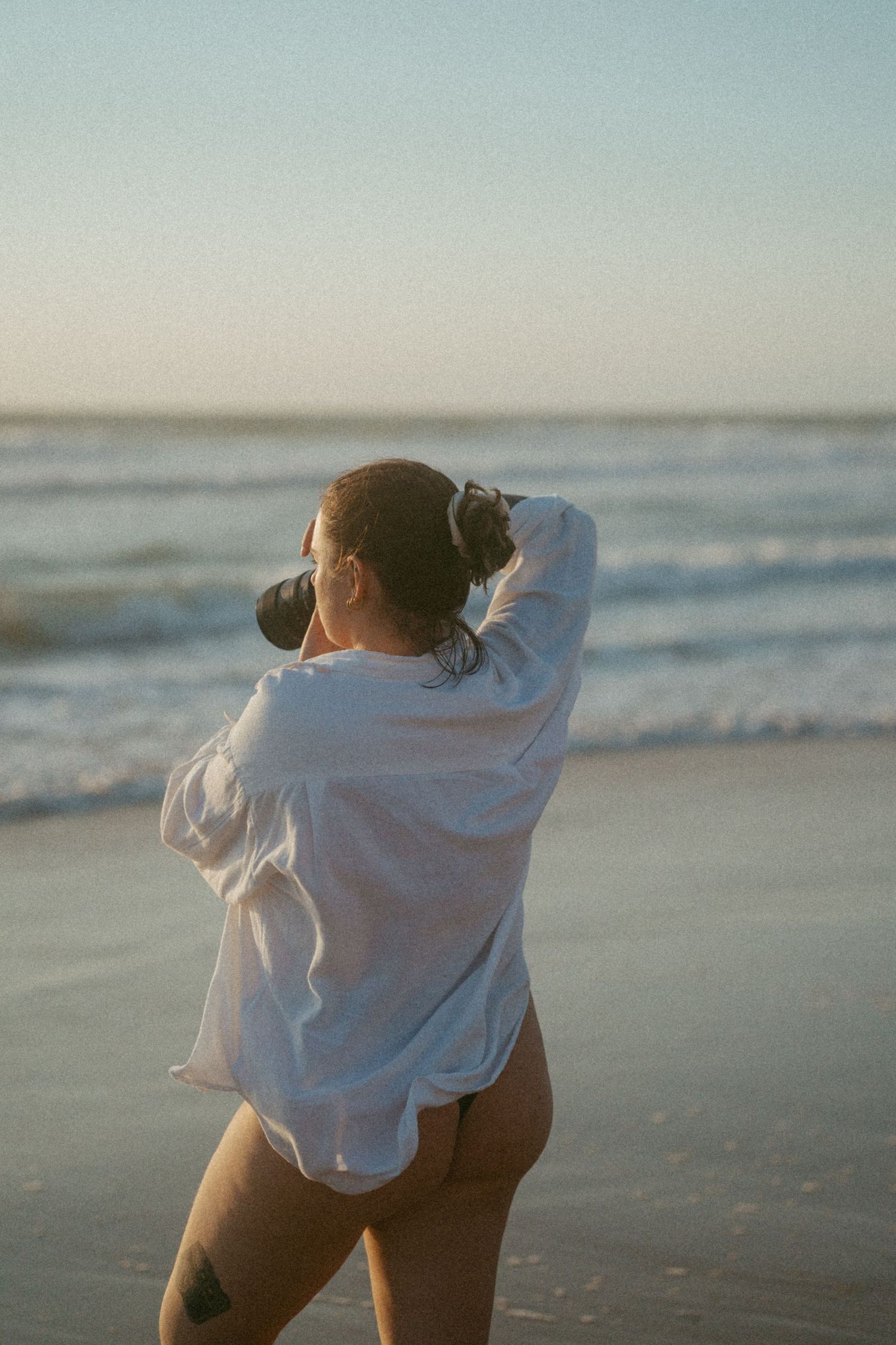A figure in an oversized white shirt stands on a beach at sunset.