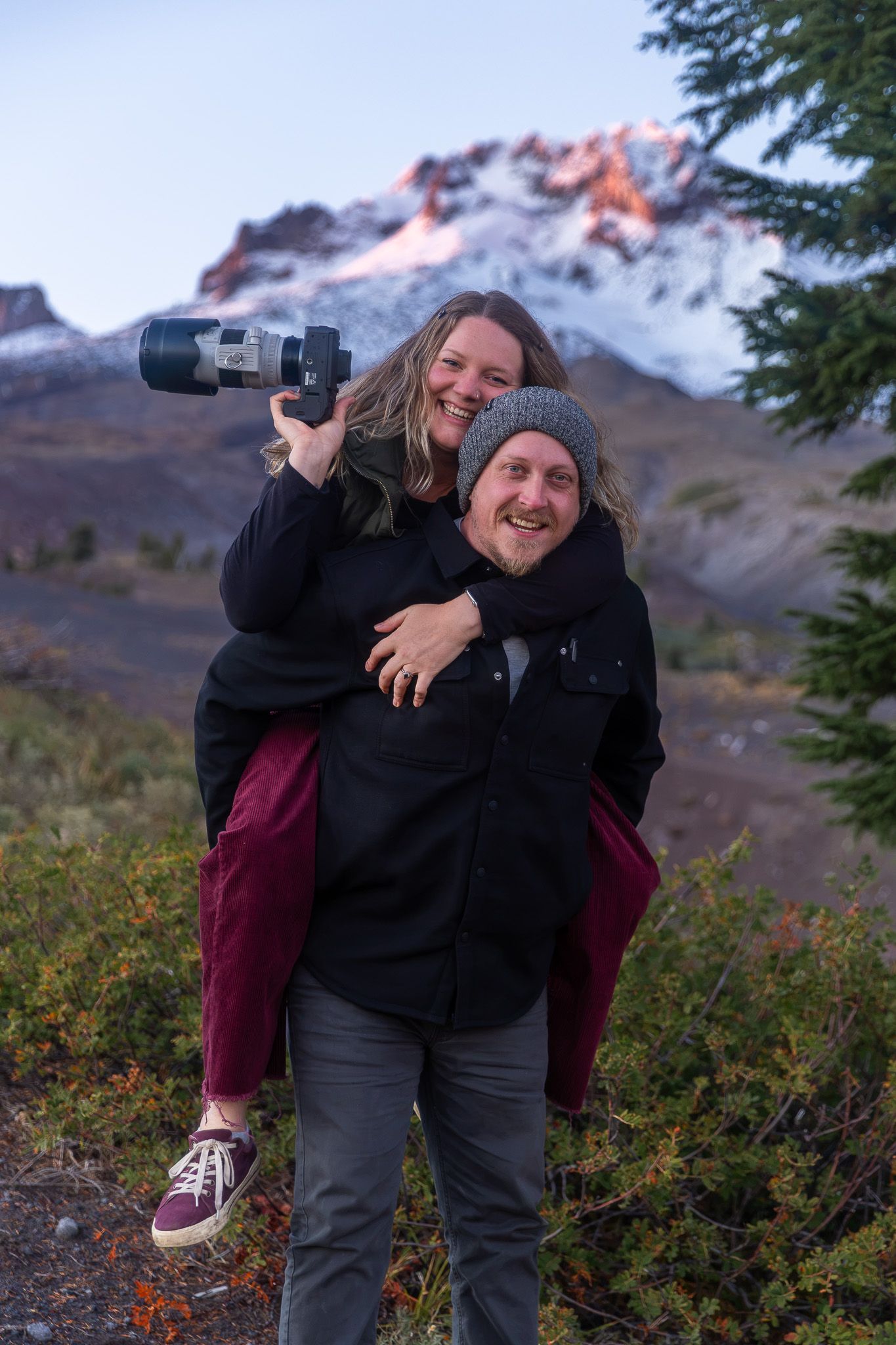 A couple smiles outdoors, woman holding camera on man's back, with snow-capped mountain and evergreen trees behind them.