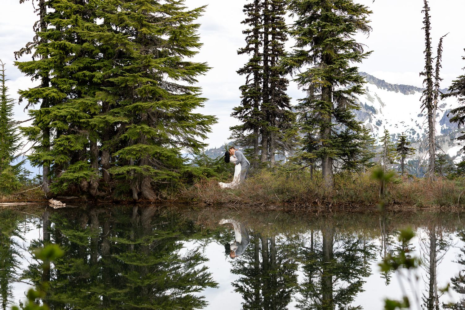 Couple kissing near a reflective alpine lake surrounded by tall evergreen trees.