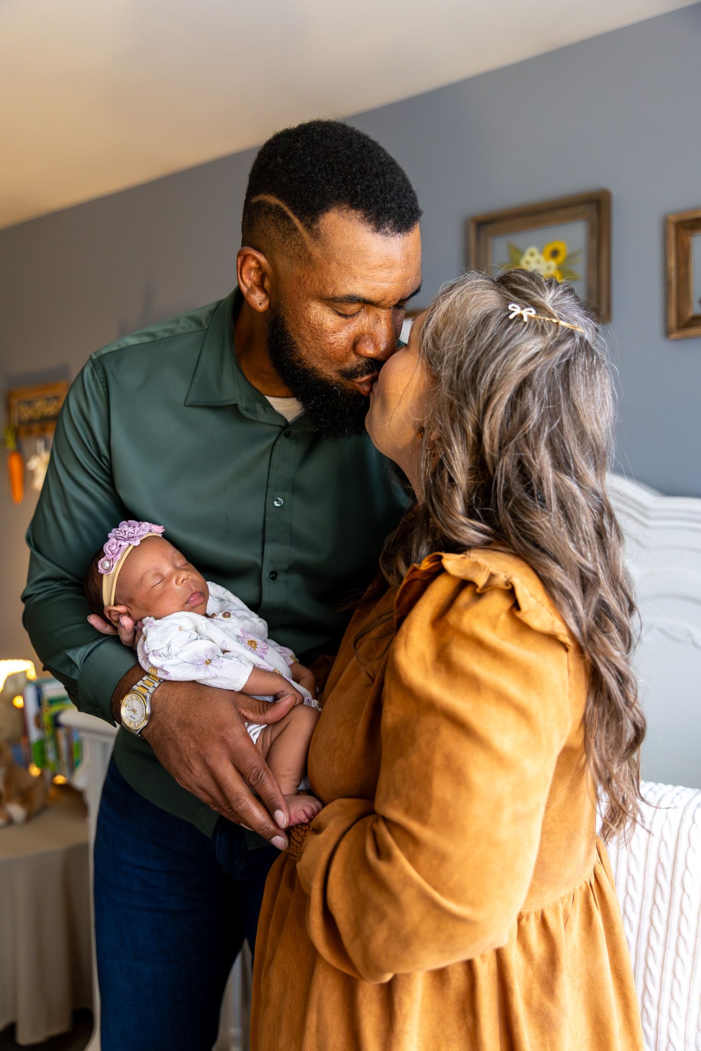 A couple shares a tender kiss while cradling their newborn baby dressed in white with a floral headband.