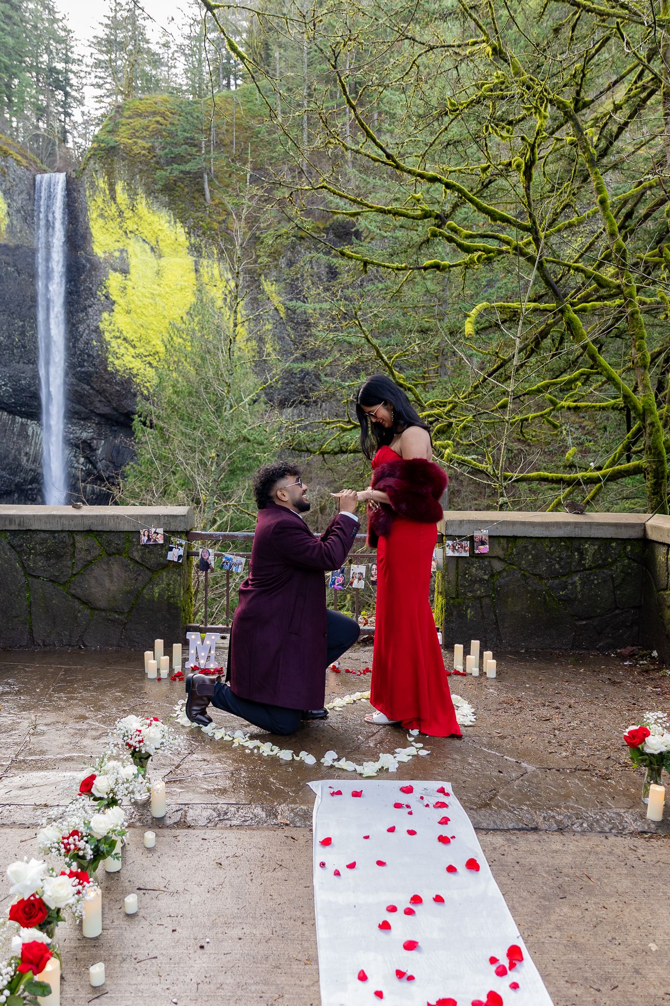 Man proposes to woman in red dress near waterfall with rose petals and candles in lush green forest setting.