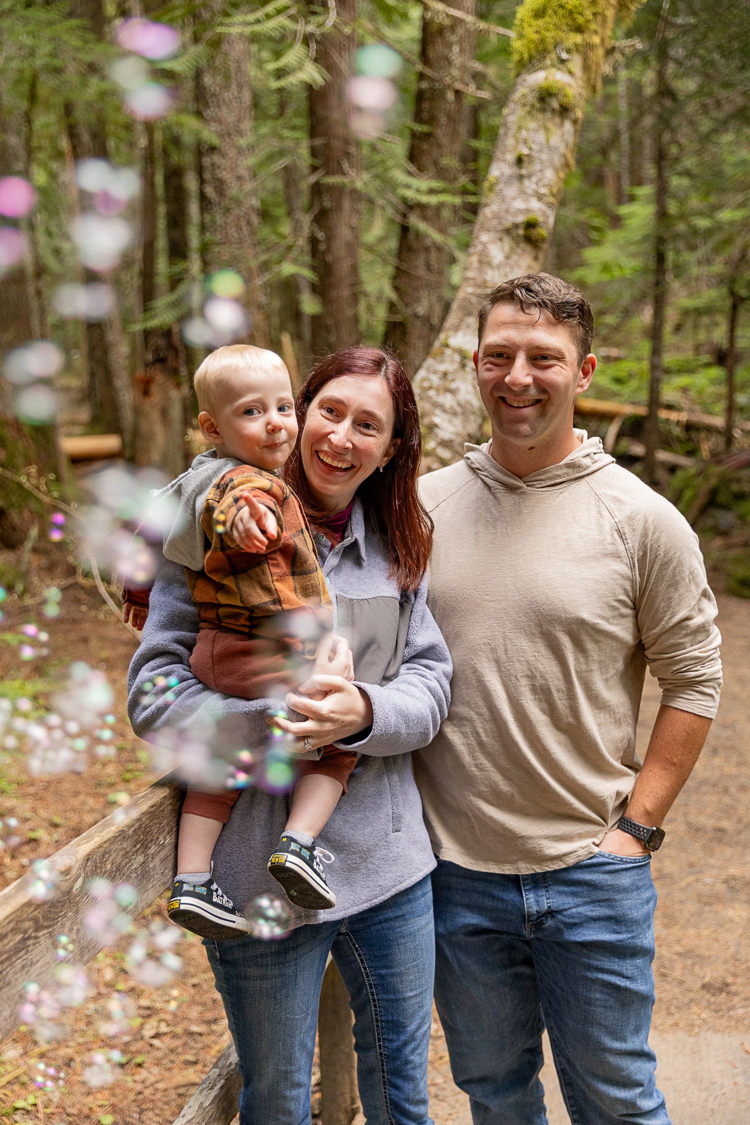 Happy family with toddler posing among bubbles on forest trail.