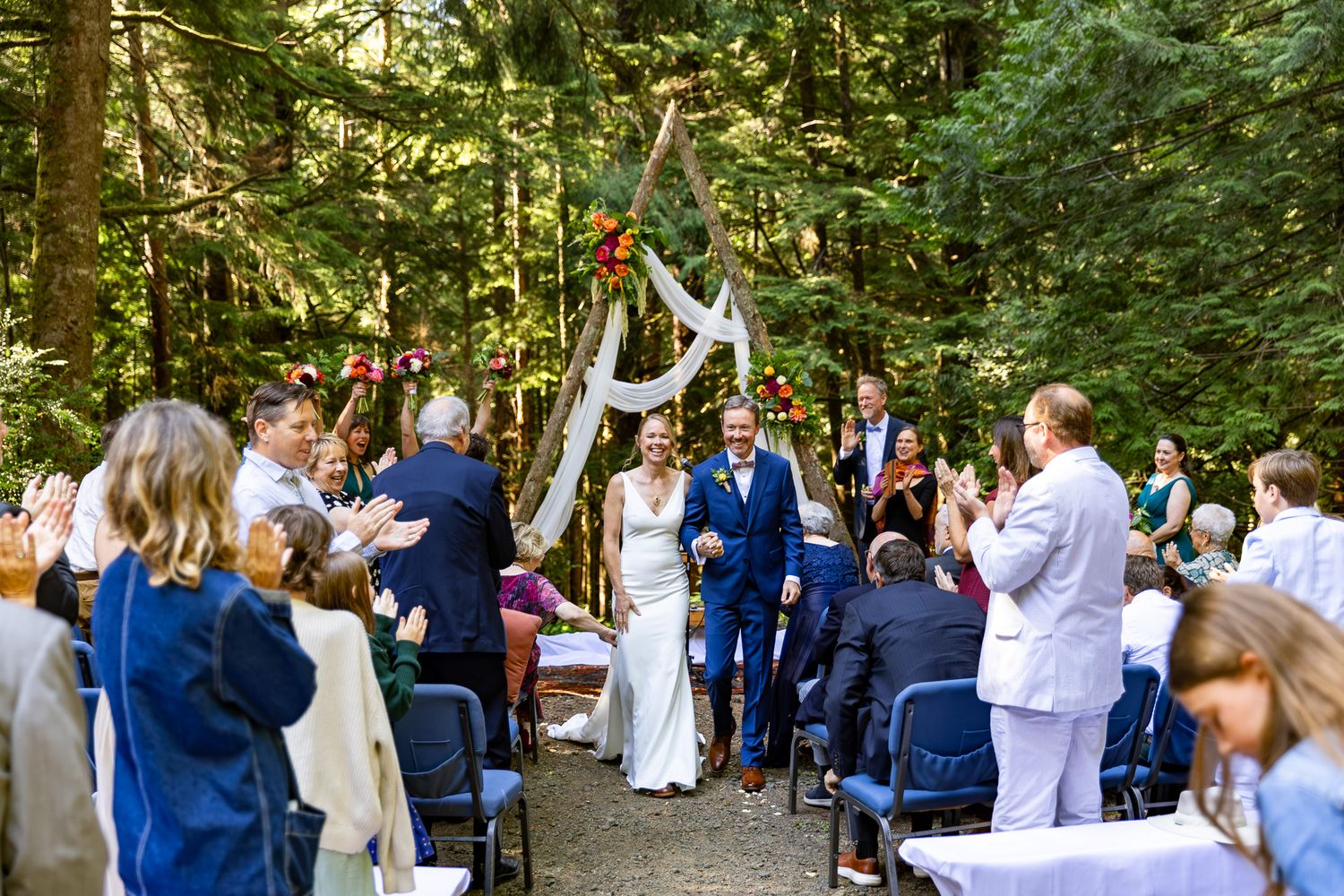 Newlywed couple walks down outdoor forest wedding aisle surrounded by applauding guests and a floral arch backdrop.