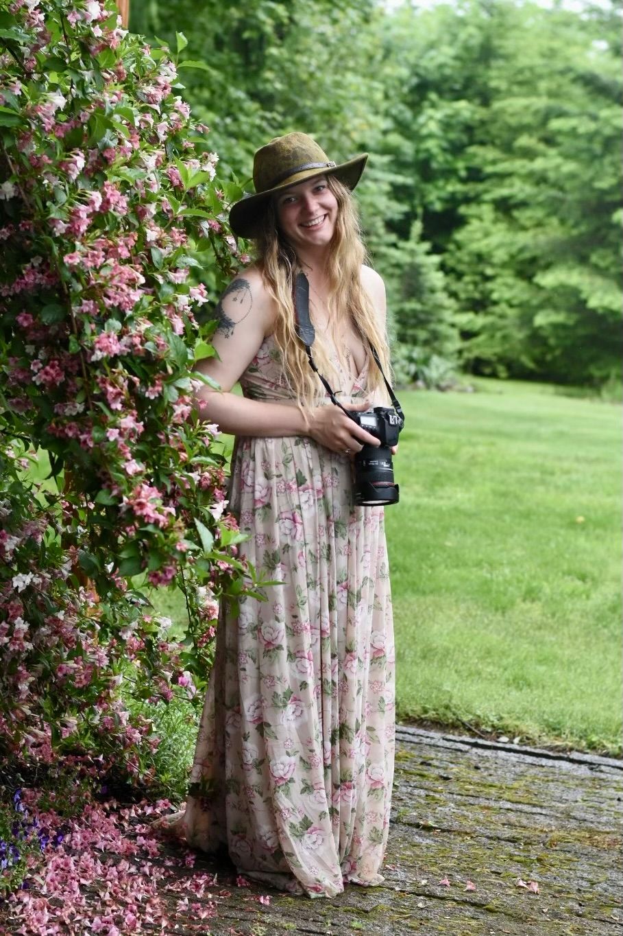 Smiling woman in floral maxi dress and wide-brim hat holding a camera beside pink flowering vines in a lush garden.