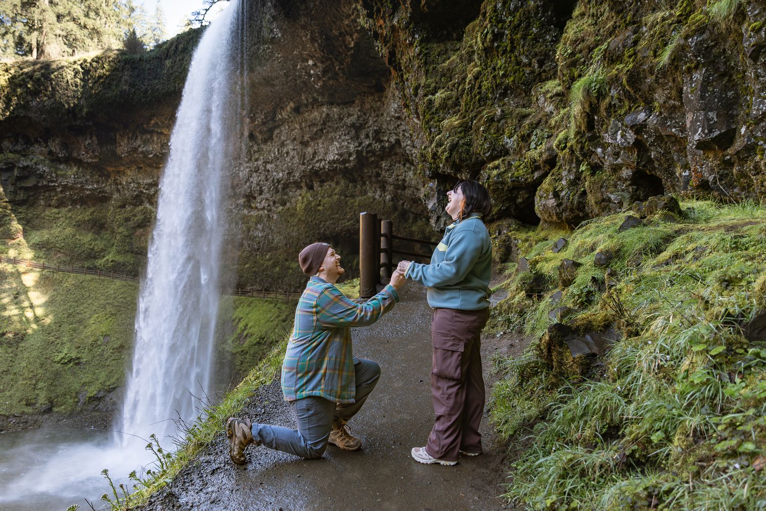 Couple sharing a romantic marriage proposal moment in front of a stunning waterfall surrounded by lush green moss.