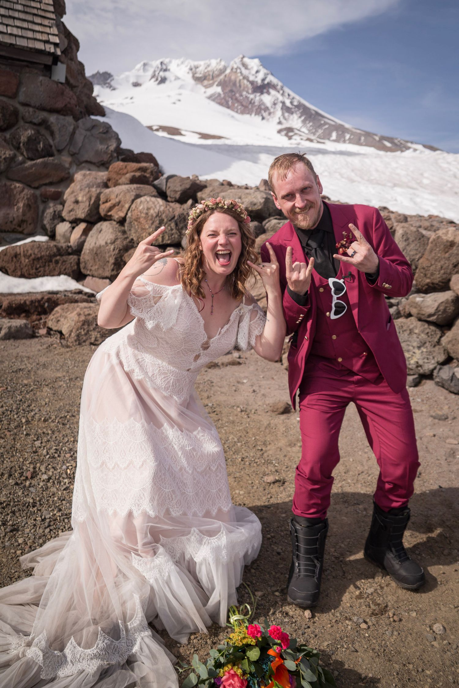 Bride and groom celebrate wedding on snowy Mt. Hood, flashing rock signs in white dress and red suit.