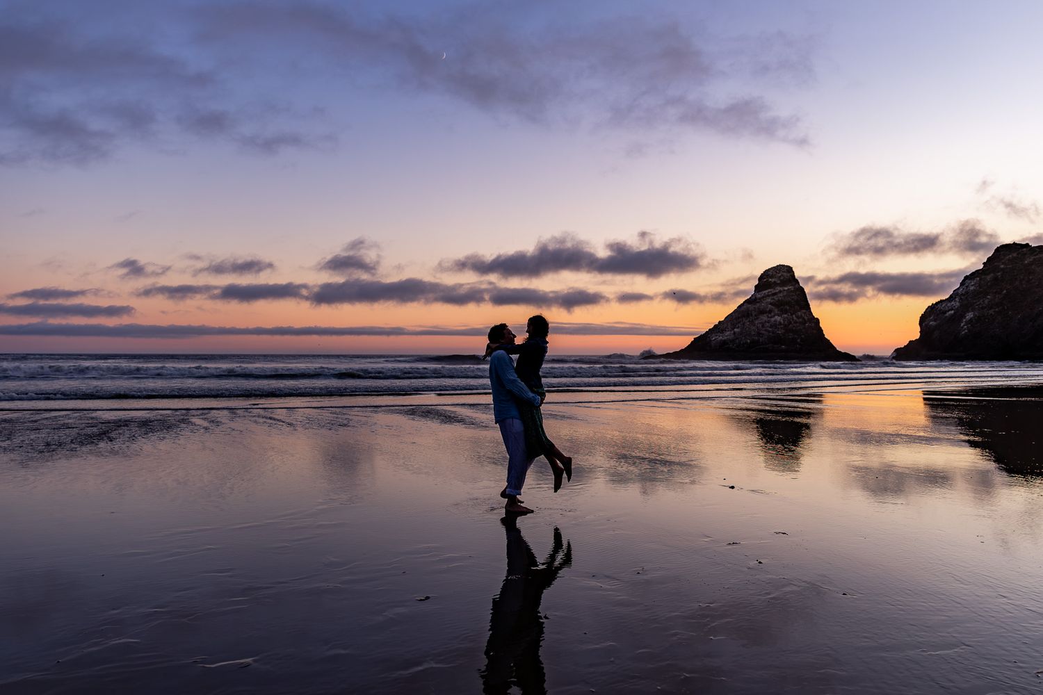 Couple silhouetted at sunset on a reflective Oregon Coast beach with dramatic rock formations and colorful sky in background.