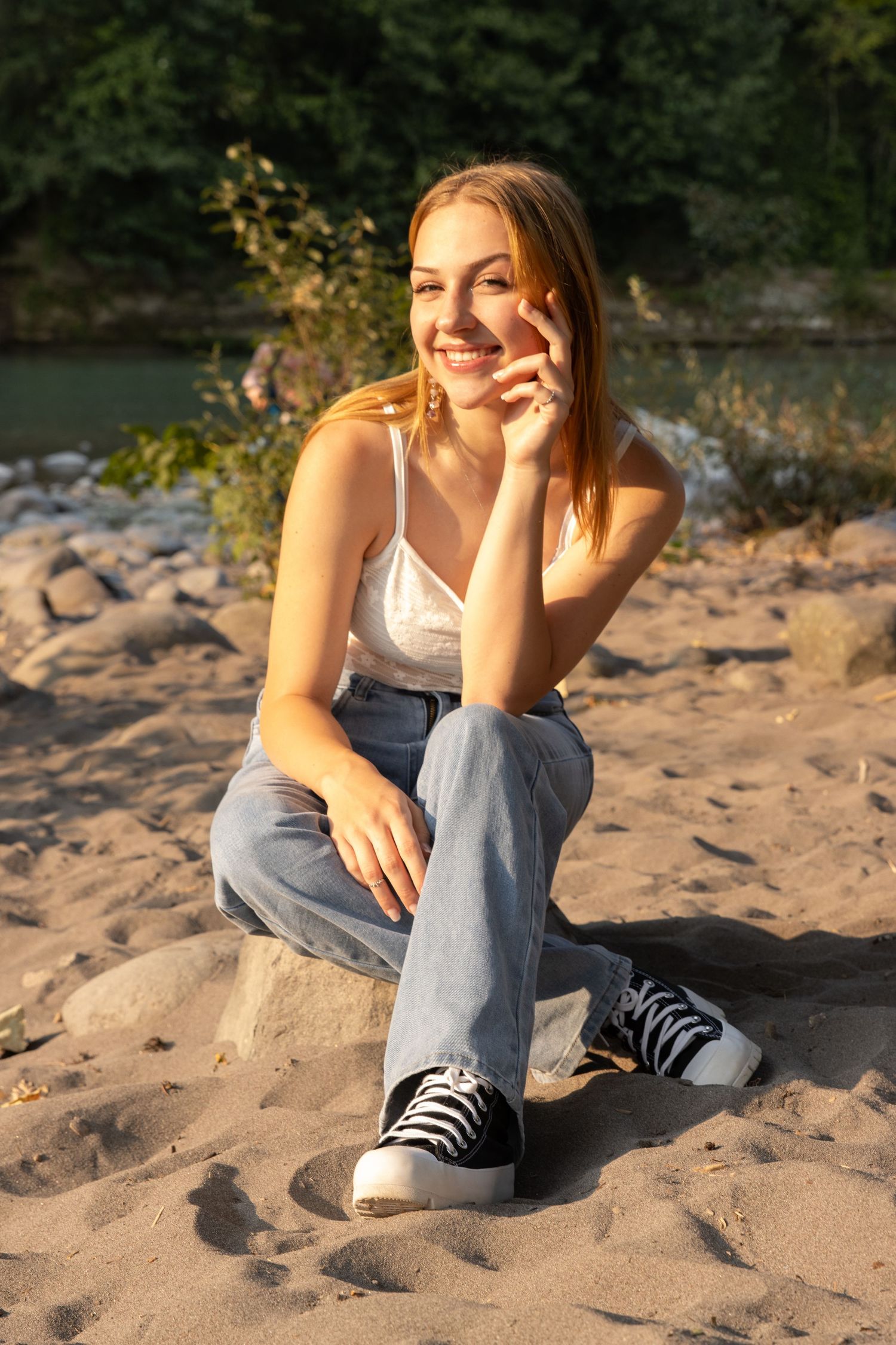 Young woman smiling in golden sunlight, sitting on a sandy riverbank in jeans and sneakers.