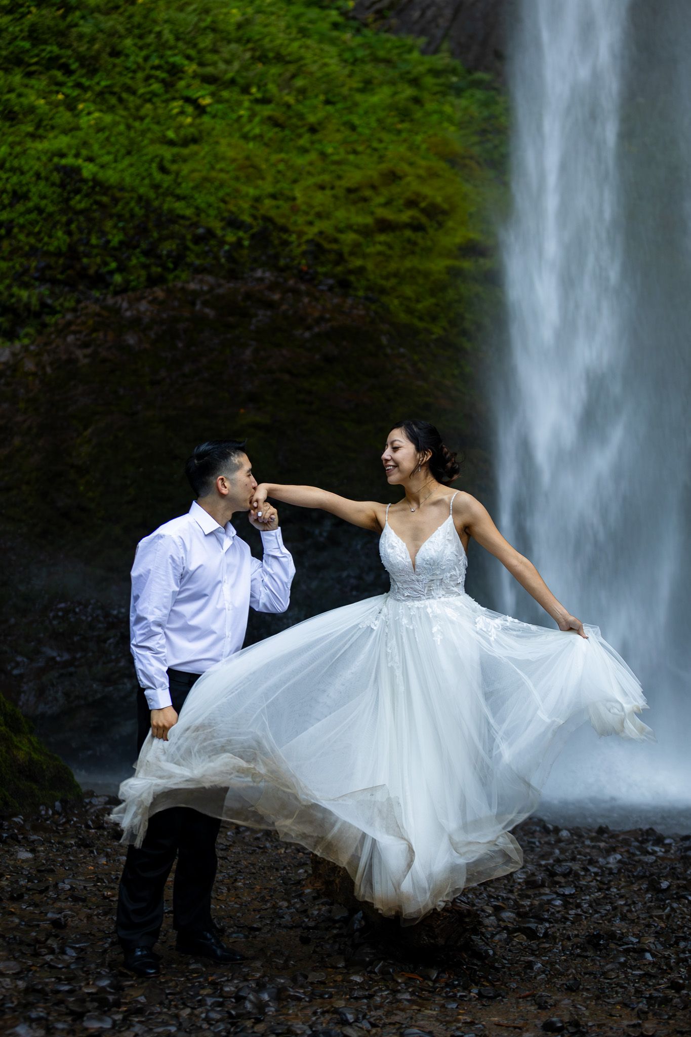 Bride and groom pose romantically at Latourell Falls. he kisses her hand as she lifts her white gown.