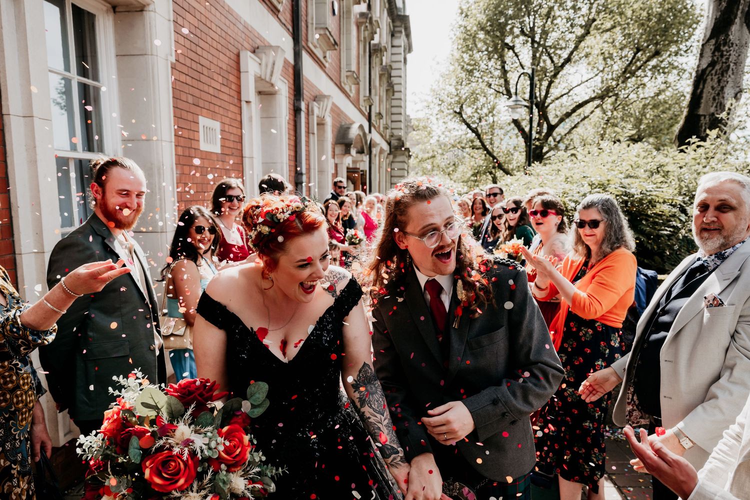 Joyful couple walks through confetti shower surrounded by cheering guests outside brick building on their wedding day.