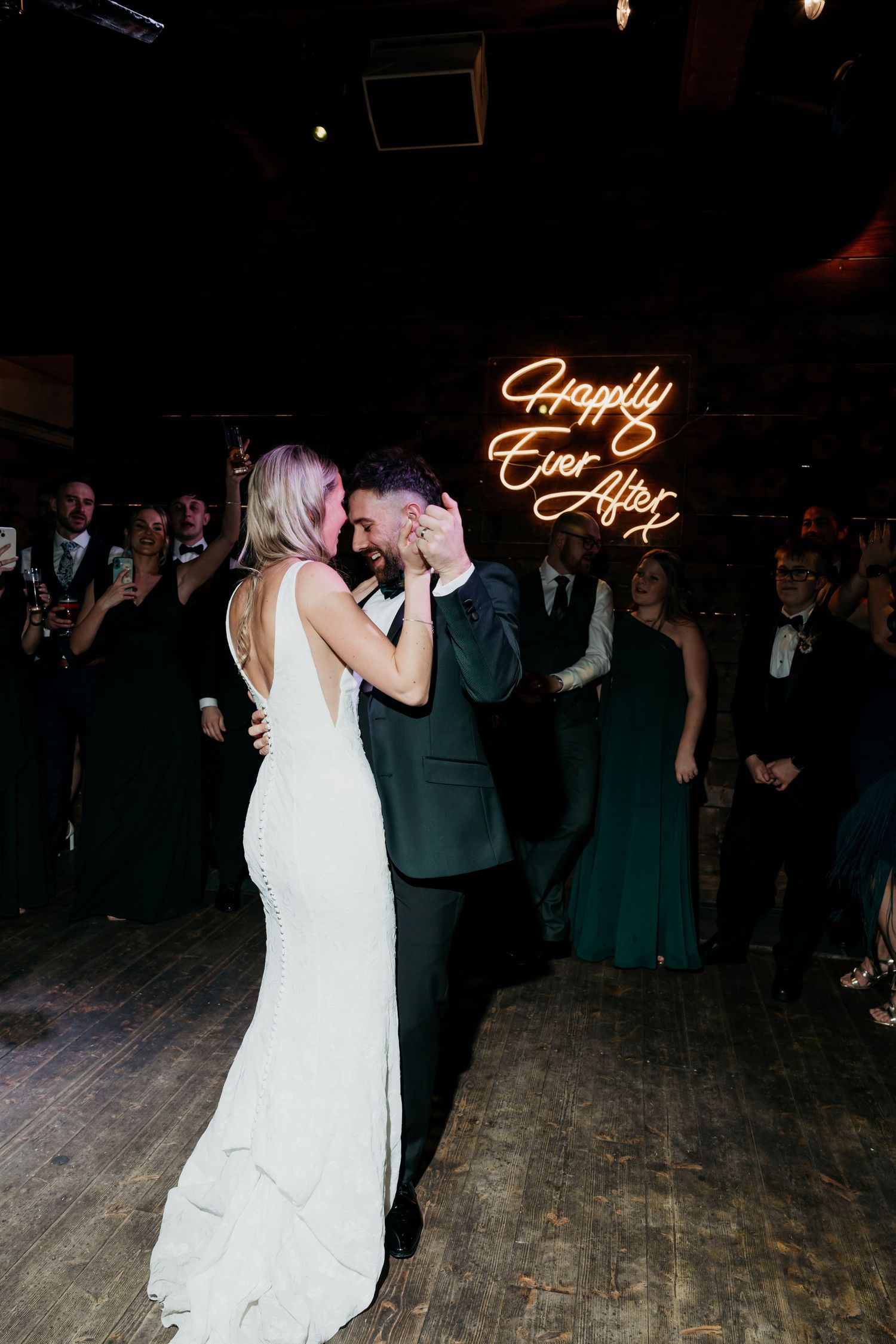 Bride and groom share first dance under a glowing Happily Ever After neon sign surrounded by wedding guests.