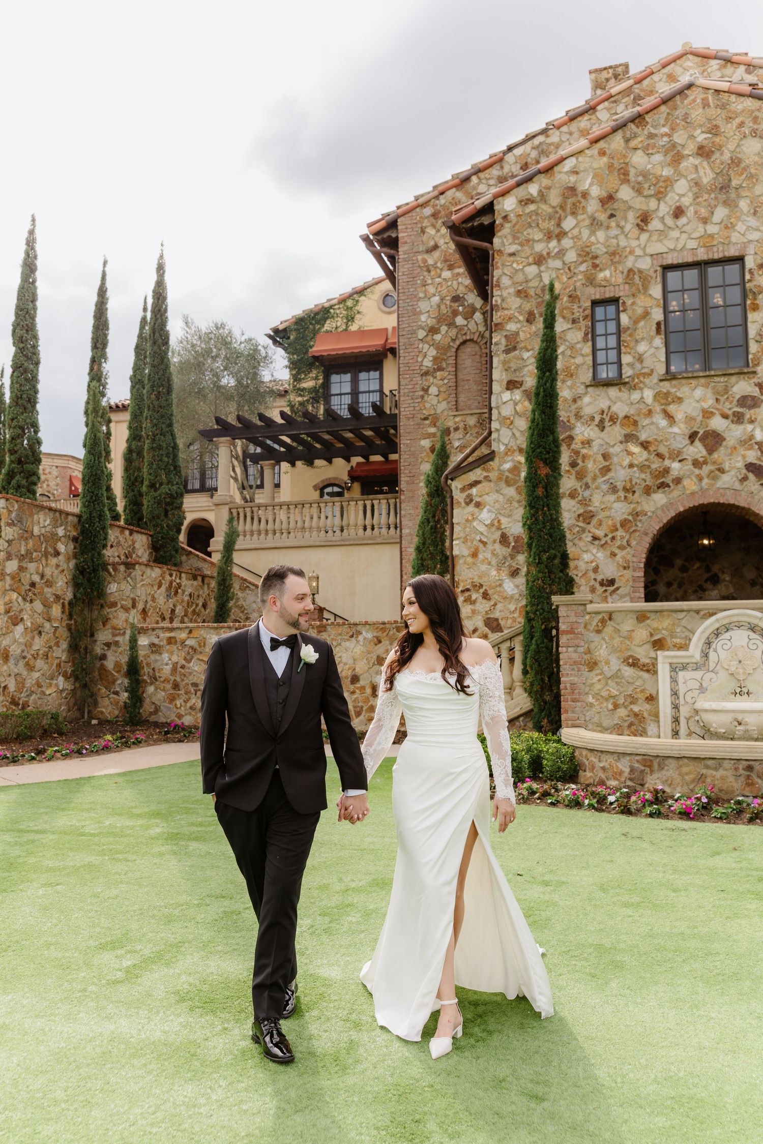 A bride and groom hold hands walking on green grass before a stone Tuscan-style villa with cypress trees.