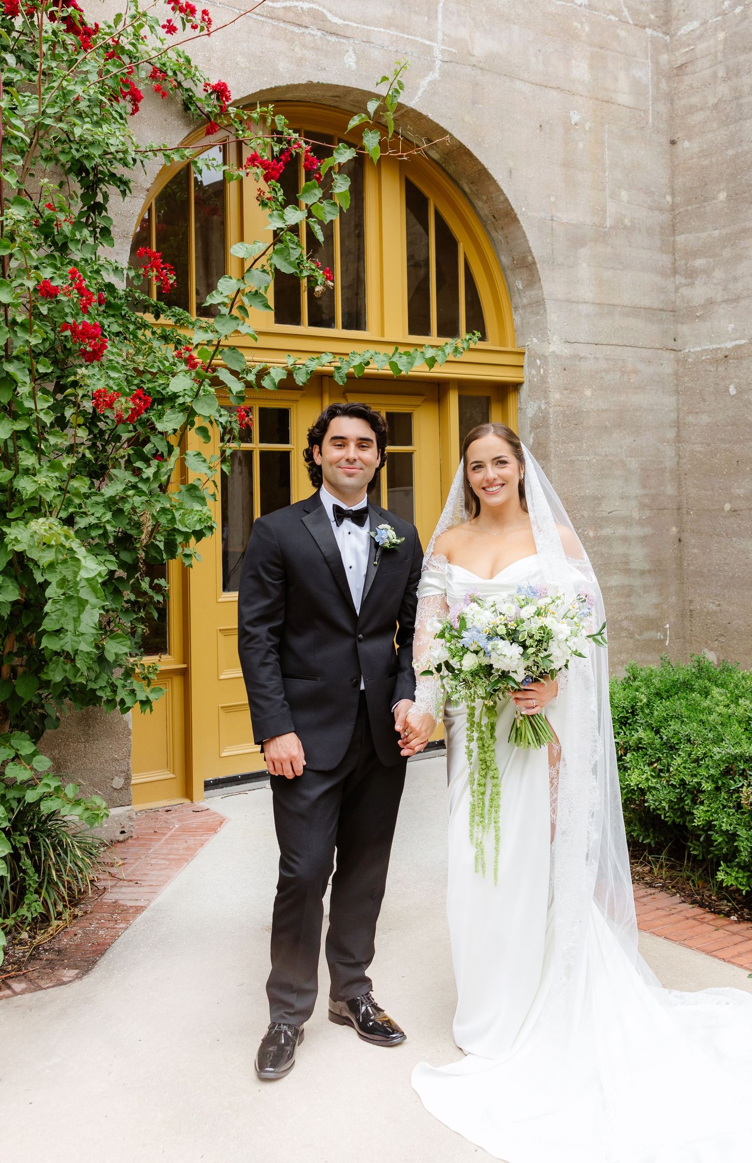 Bride and groom holding hands in front of yellow arched door with red climbing roses, bride holding cascading white bouquet.