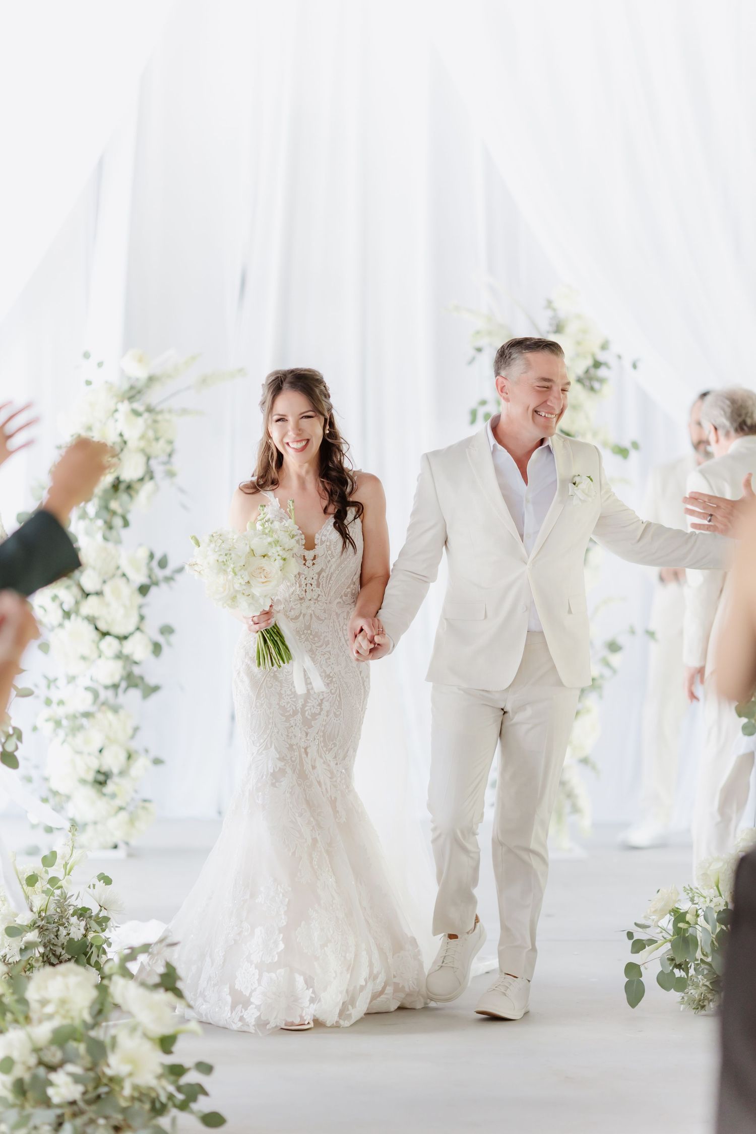 Smiling newlyweds walk down the aisle, bride in lace gown holding white bouquet, groom in cream suit, surrounded by floral arches.