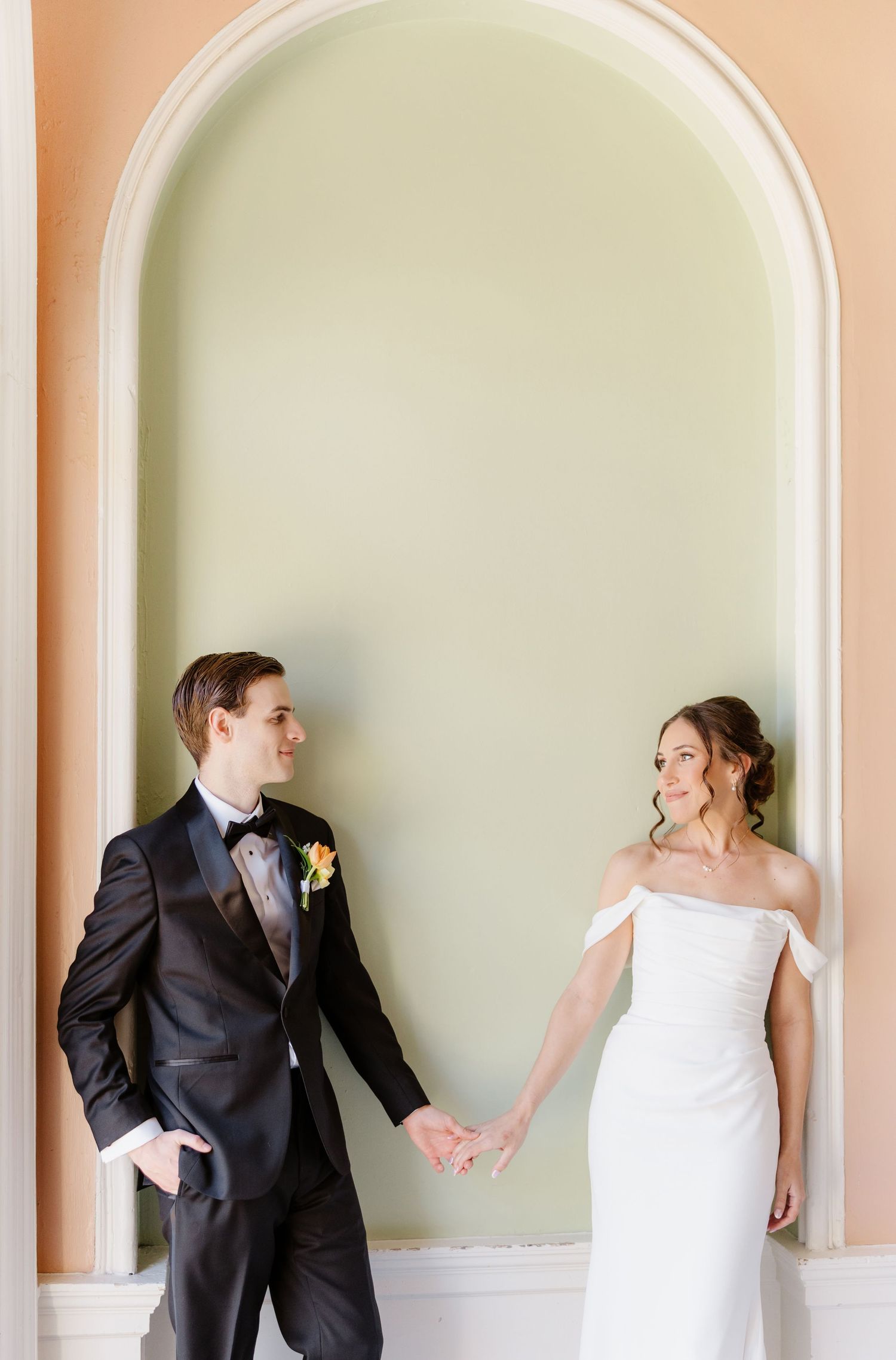 Bride and groom hold hands in front of a sage green arched alcove, bride in off-shoulder gown, groom in black tuxedo.