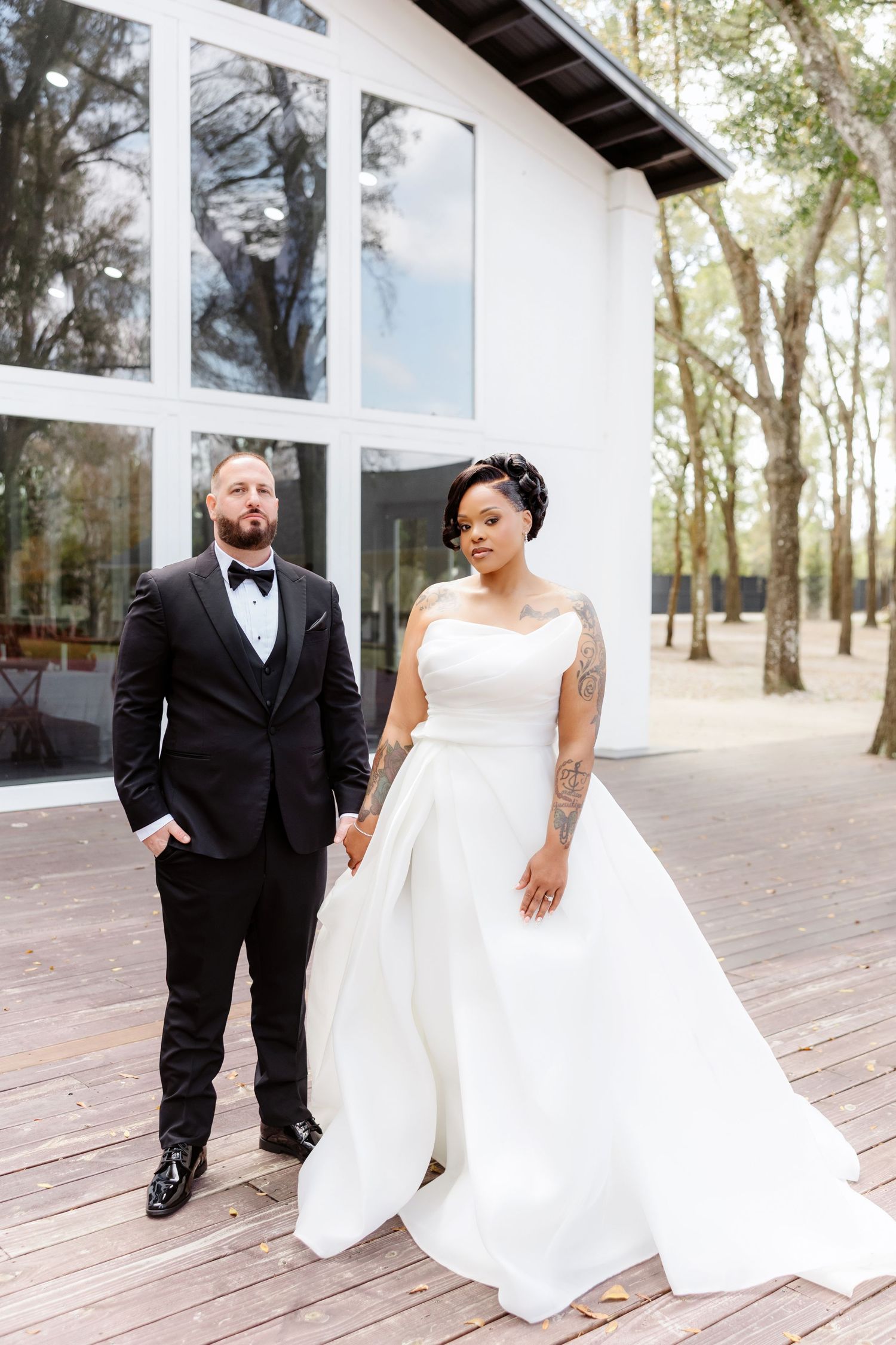 Bride and groom in formal wedding attire stand on wooden deck outside modern white building surrounded by trees.