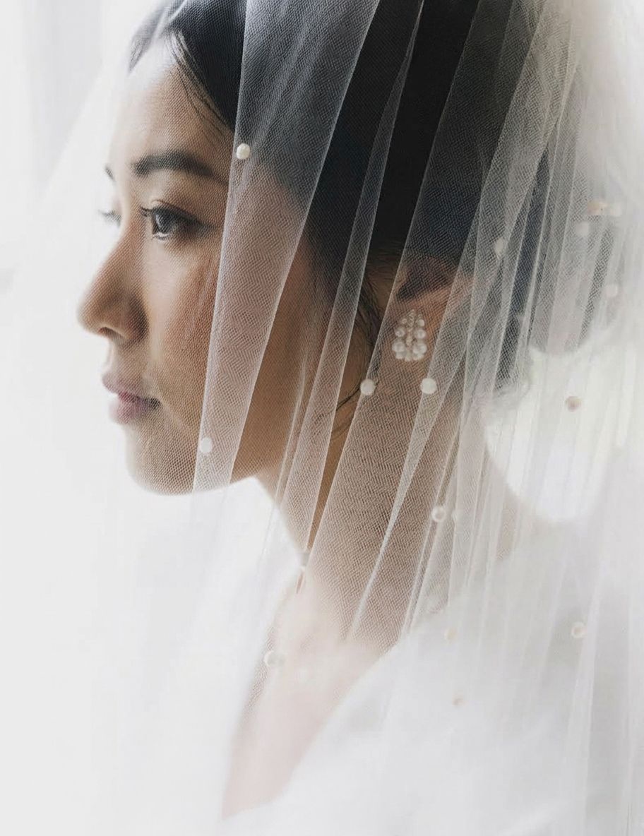 A bride in profile wearing a delicate pearl-adorned veil and floral earring, with soft natural light illuminating her features.