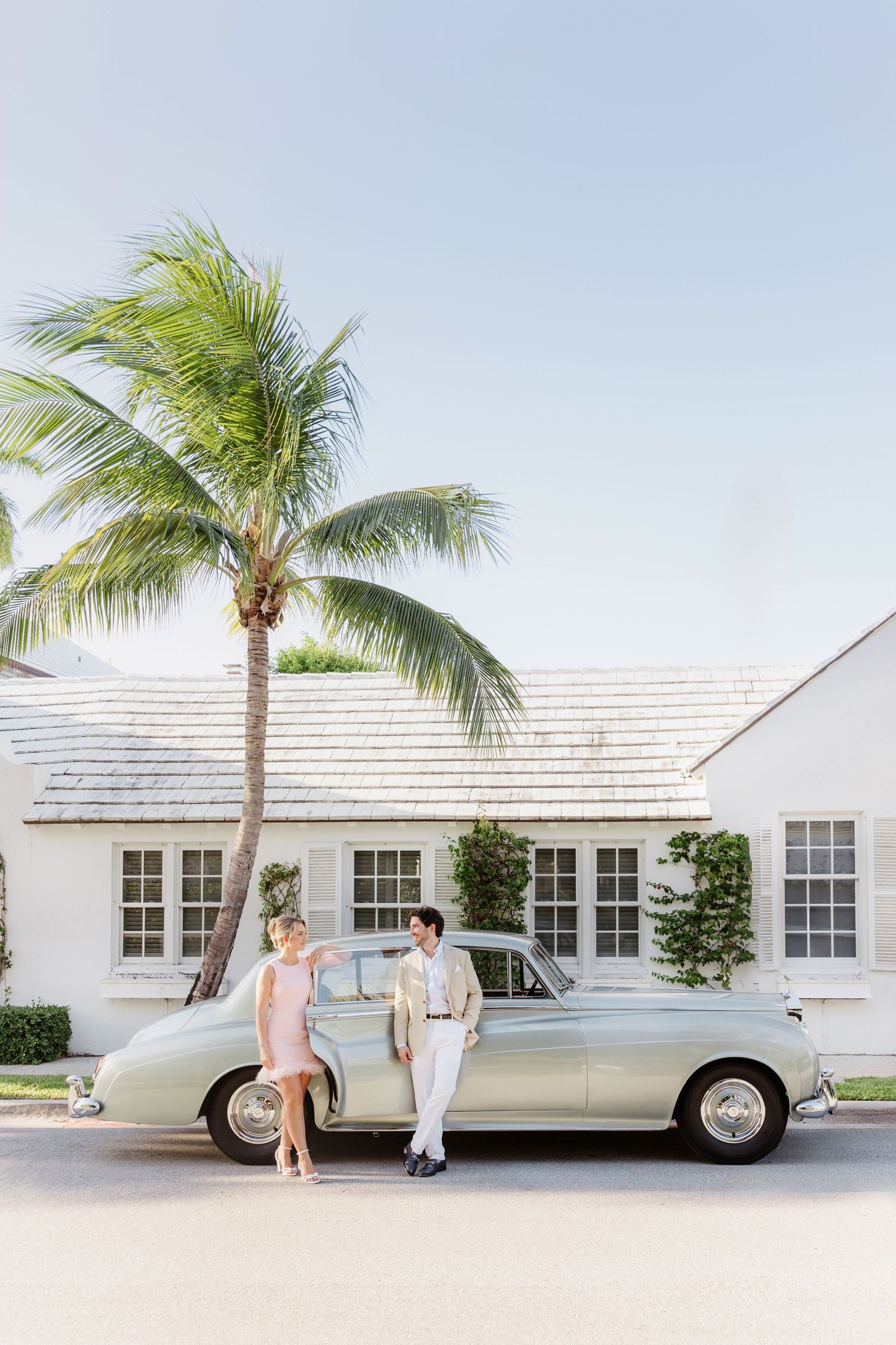 Couple posing by a vintage mint green car in front of a white house with a palm tree in a sunny tropical setting.