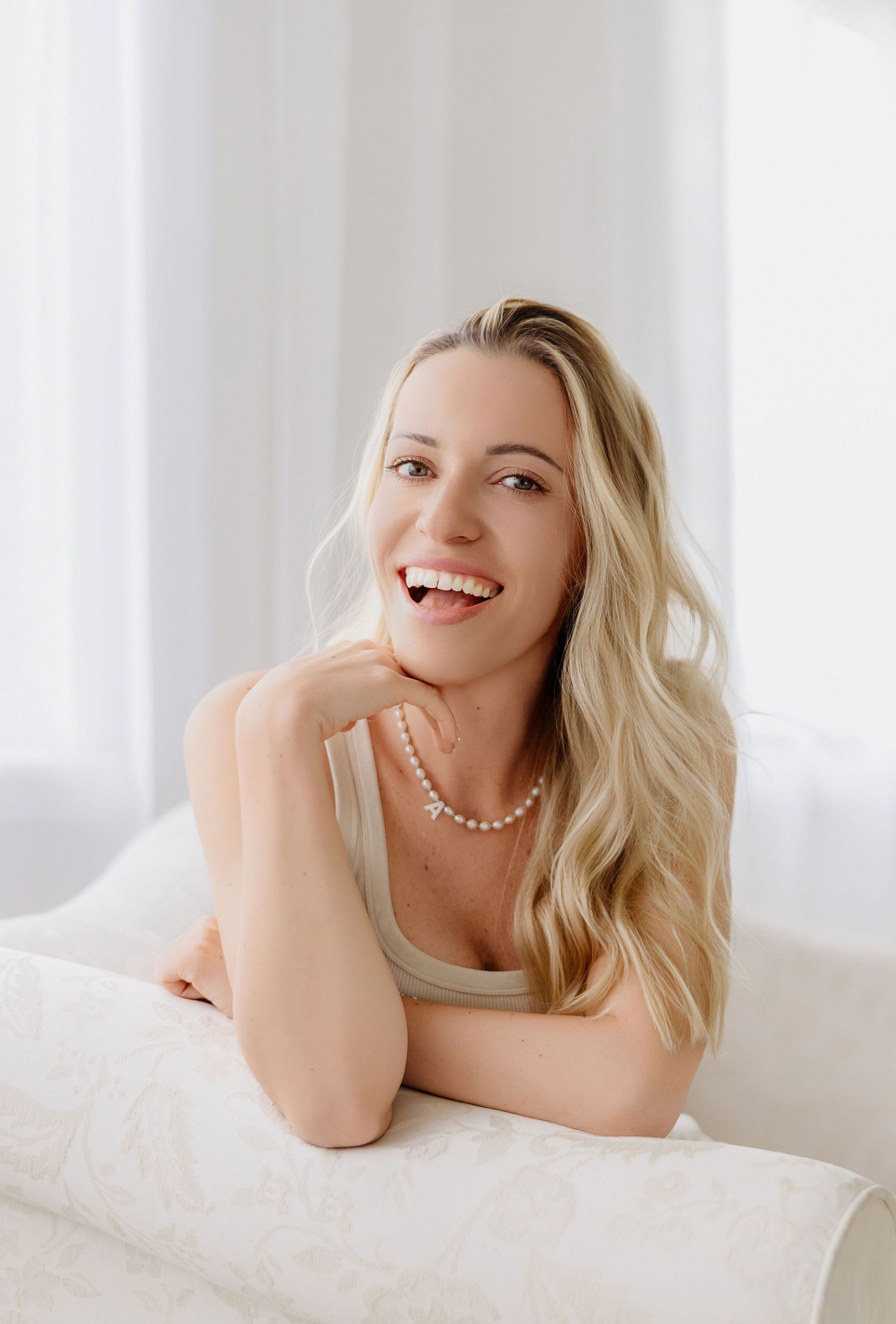 Smiling blonde woman with pearl necklace leaning on white sofa in bright airy room.