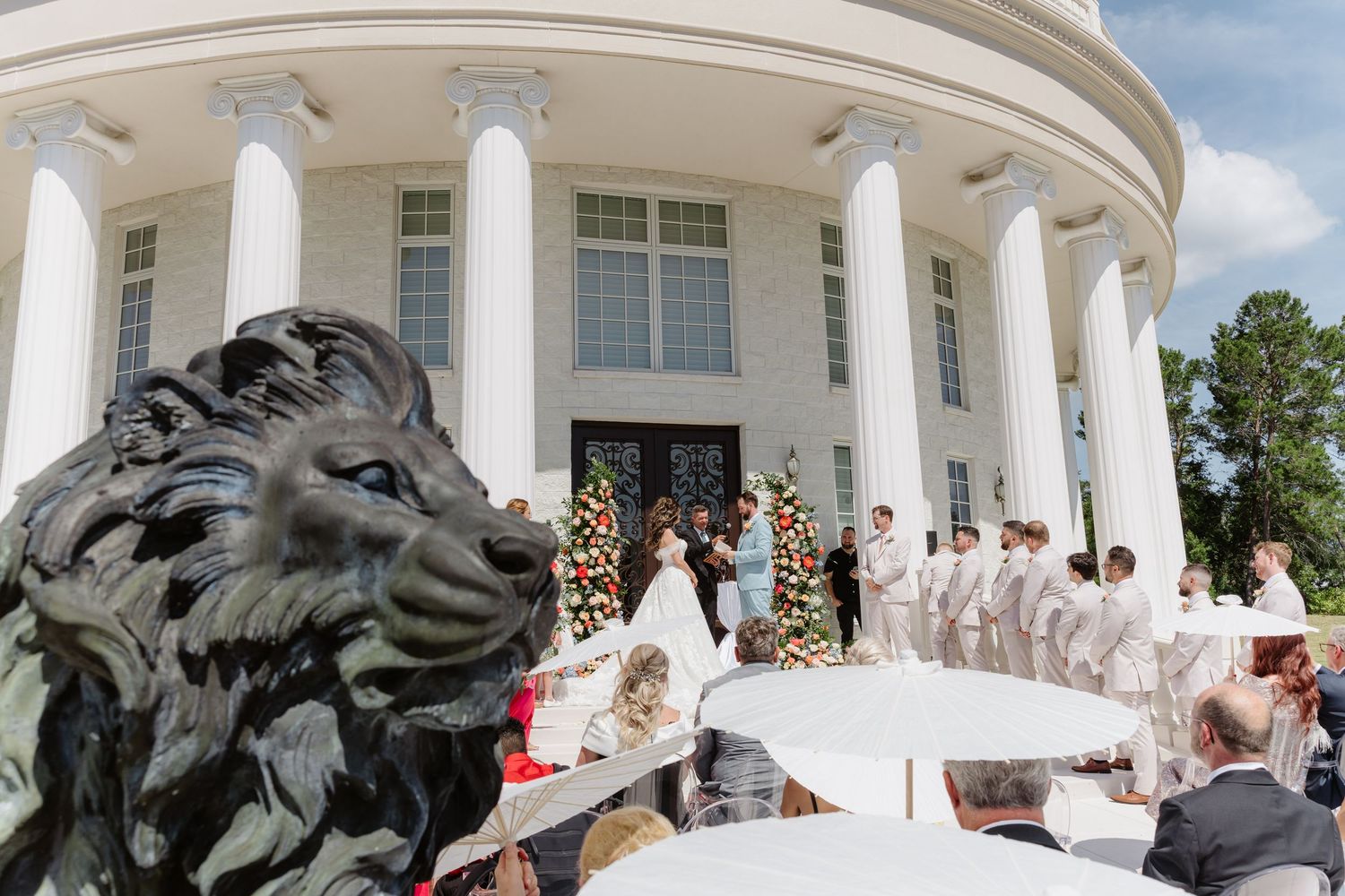 Outdoor wedding ceremony at a white columned mansion with bridal party, floral arch, and bronze lion statue in foreground.