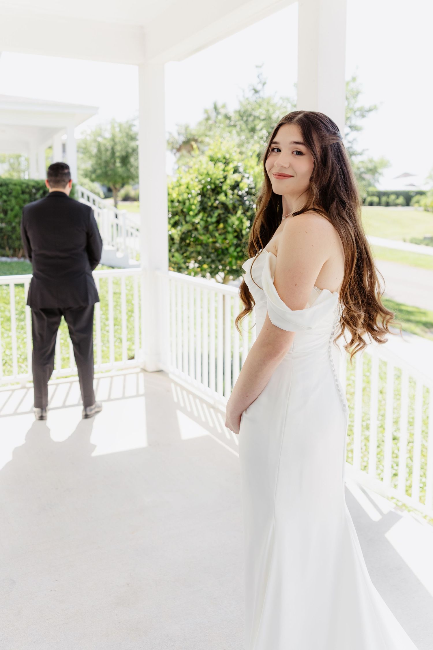 Bride in white off-shoulder gown smiles on porch while groom in dark suit stands facing away.