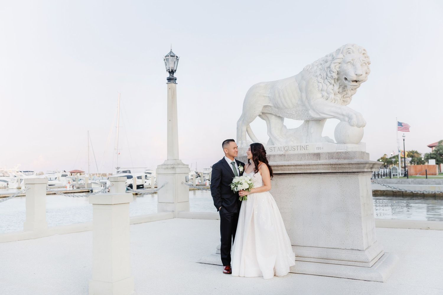 Bride and groom pose beside a white lion statue on a waterfront terrace during their elegant outdoor wedding ceremony.