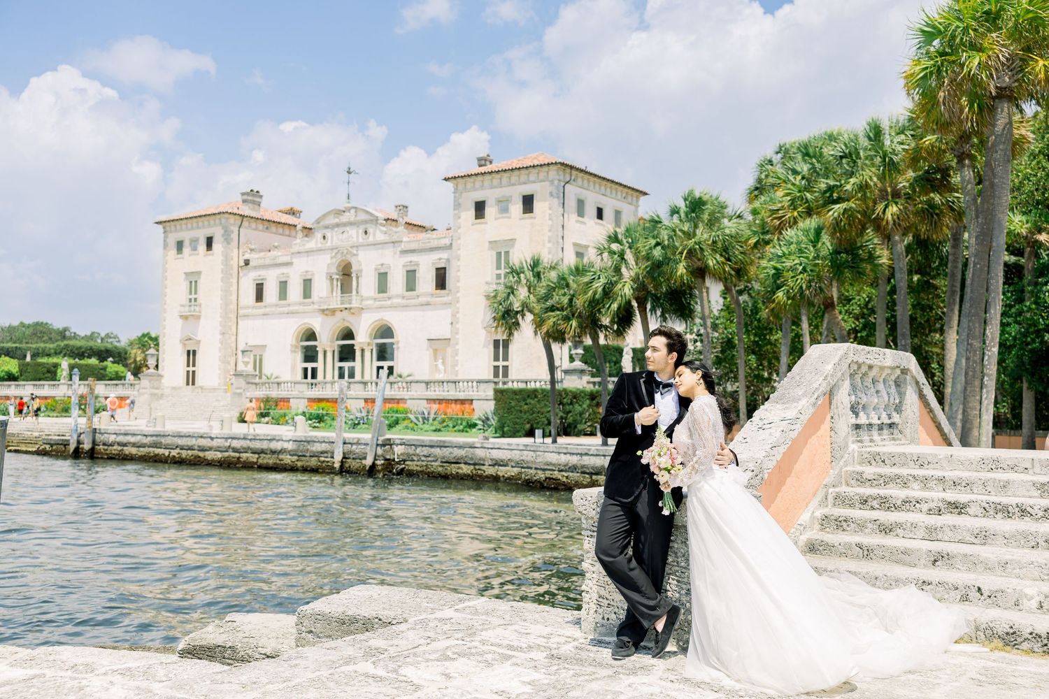 A couple in wedding attire poses at Vizcaya Museum and Gardens in Miami with palm trees and stone architecture.