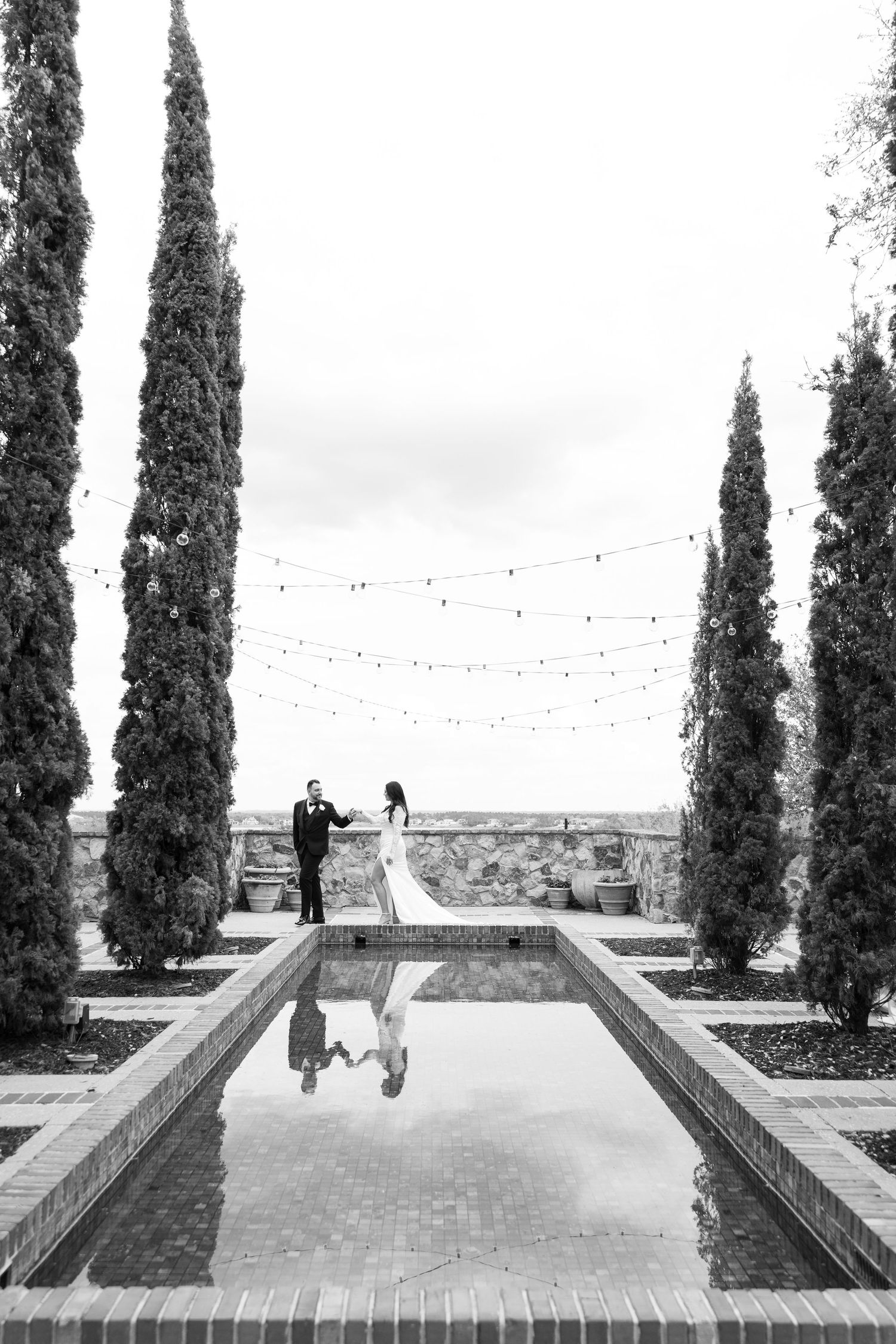 Black and white wedding photo of bride and groom holding hands by a reflecting pool lined with tall cypress trees.