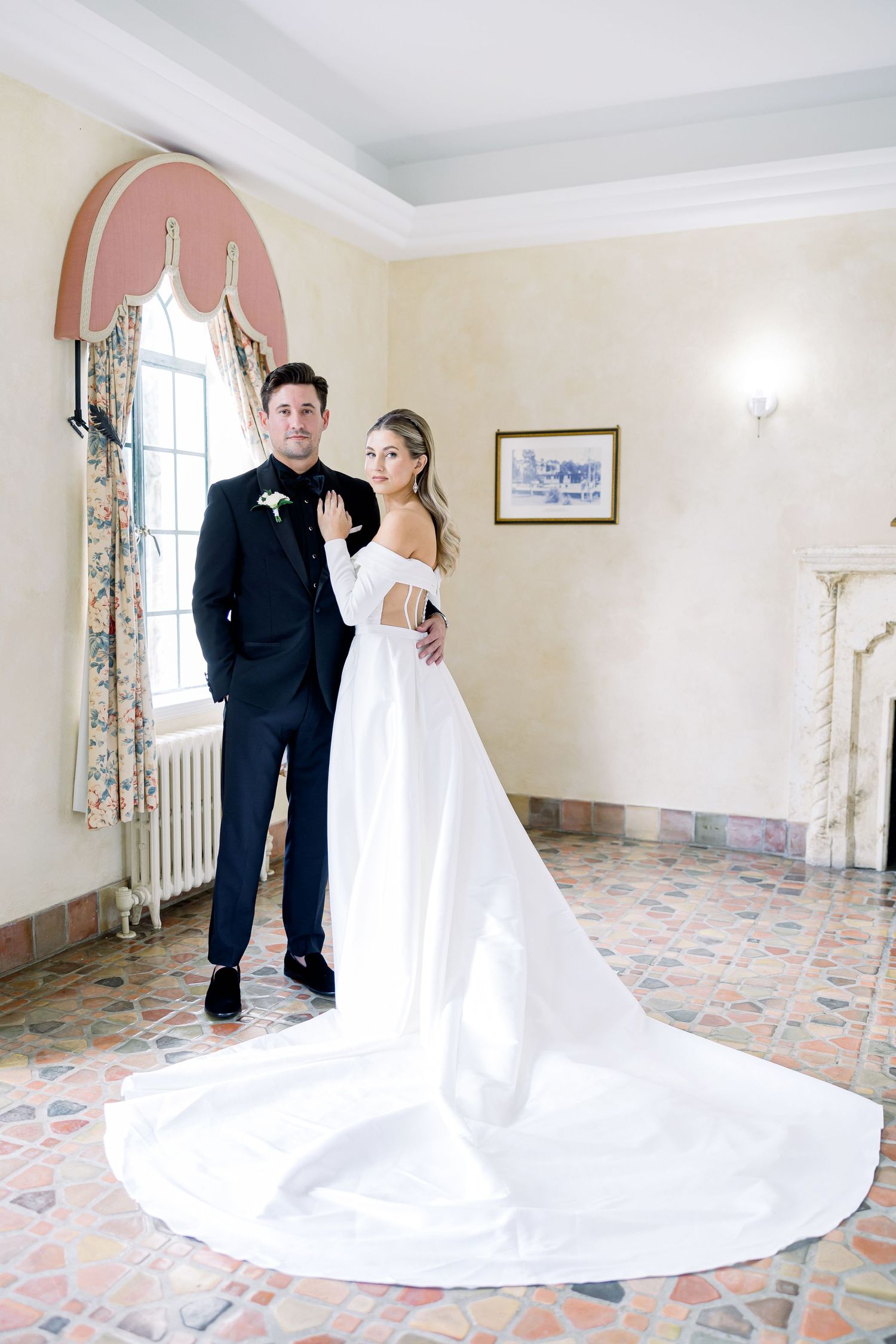 A wedding couple poses in a room with pink arched walls, white curtains and floral tile flooring.